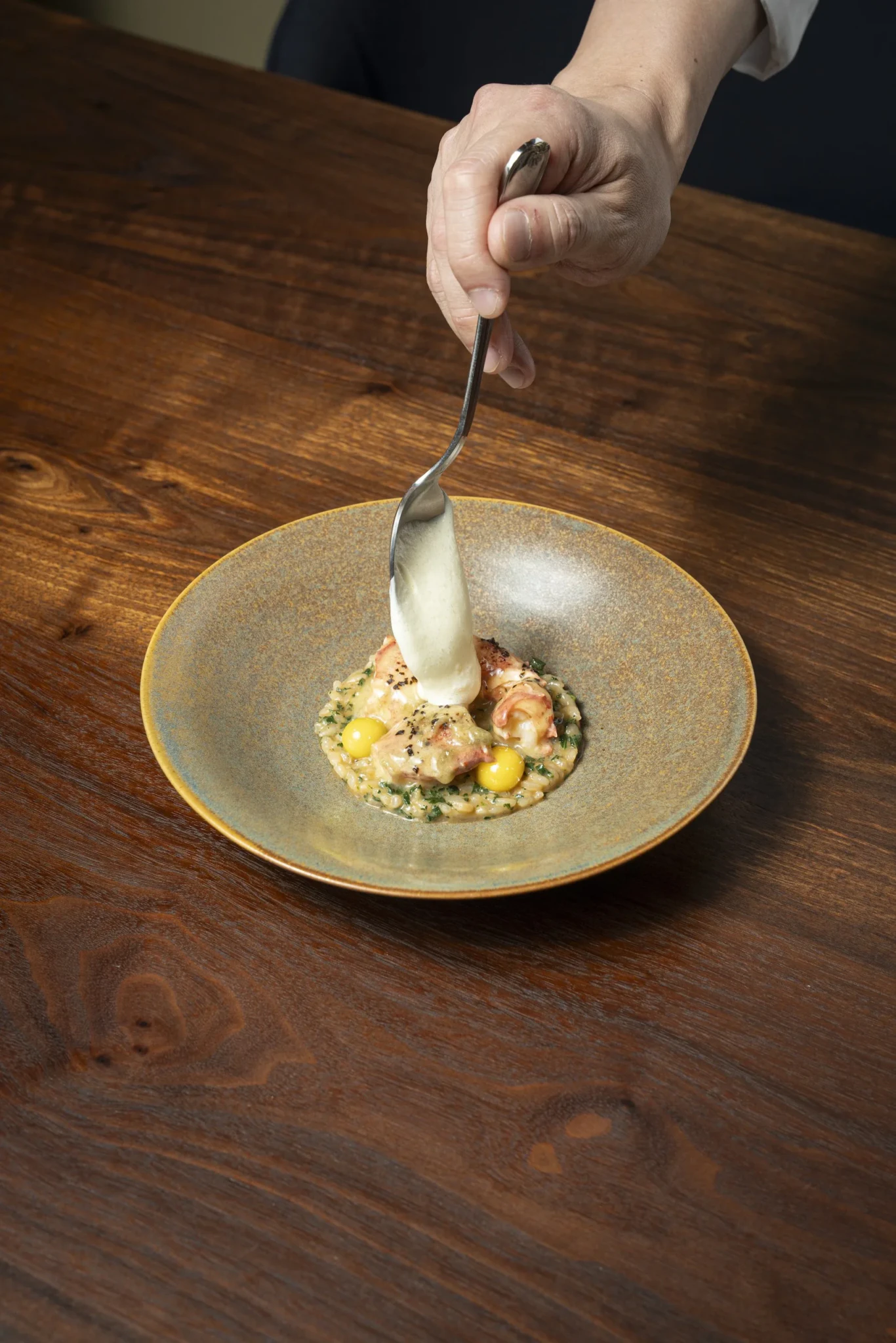 A spoon drizzling creamy sauce over lobster and herb-flecked rice in a speckled ceramic bowl at Restaurant Allen