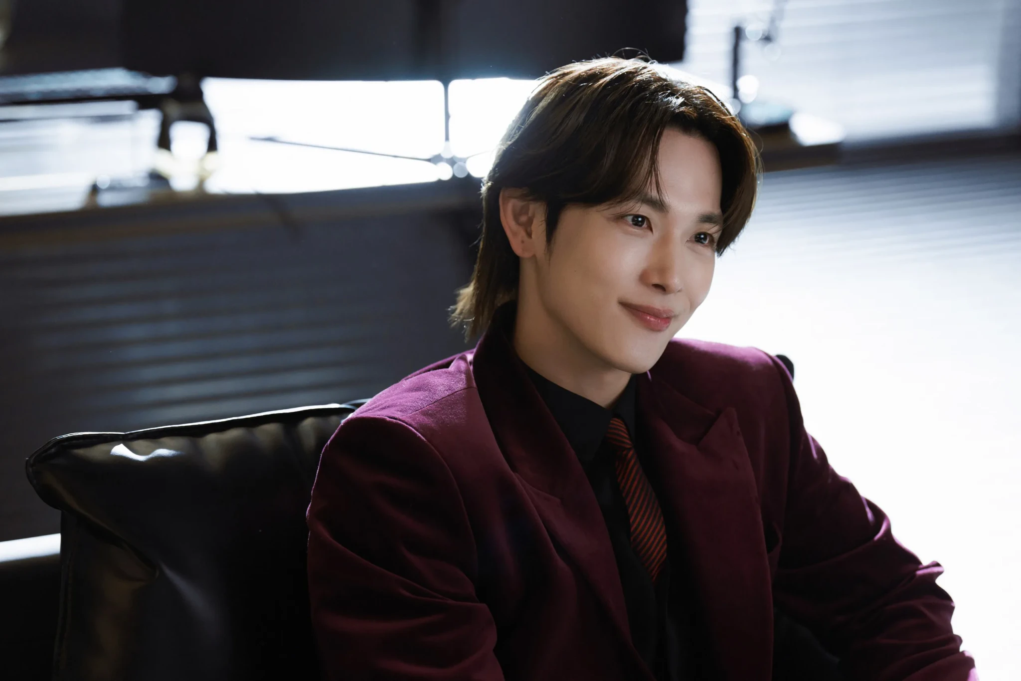 A man in a burgundy velvet suit, black shirt and striped tie smiles from a black leather chair in a Love in Disguise still