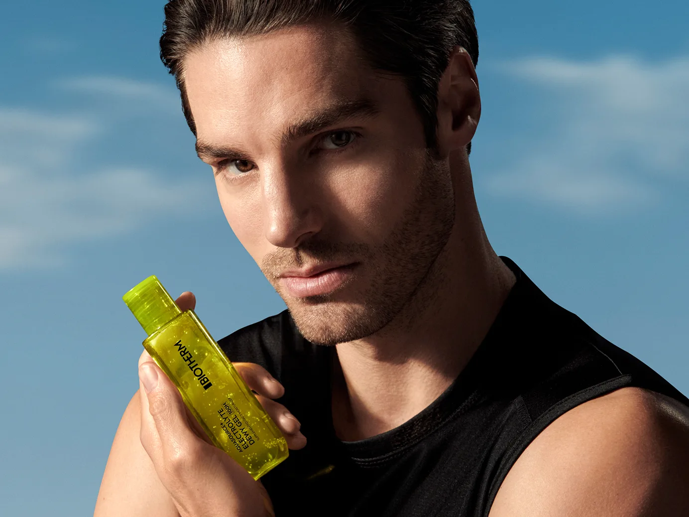 Man in a black sleeveless top holding Biotherm Aquasource+ Electrolyte Dewy Gel against a blue sky