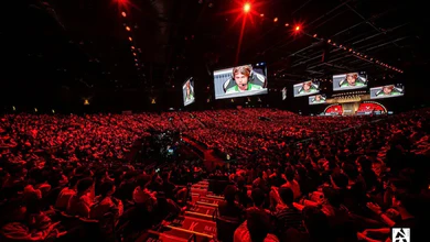 A packed crowd lit by red stage lighting, with large screens hanging above the arena floor
