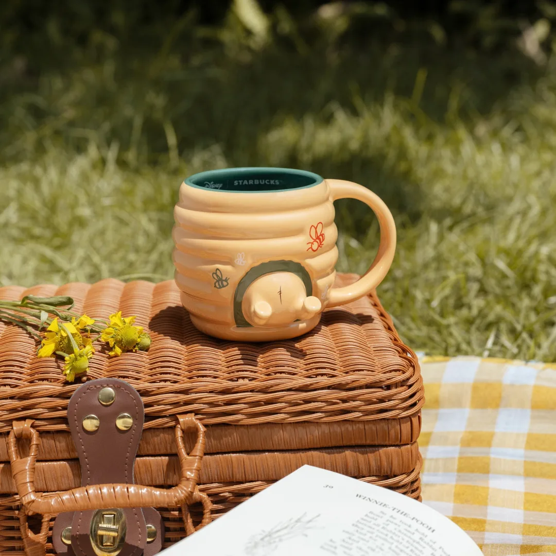 Starbucks Winnie the Pooh beehive mug on a wicker picnic basket beside an open book