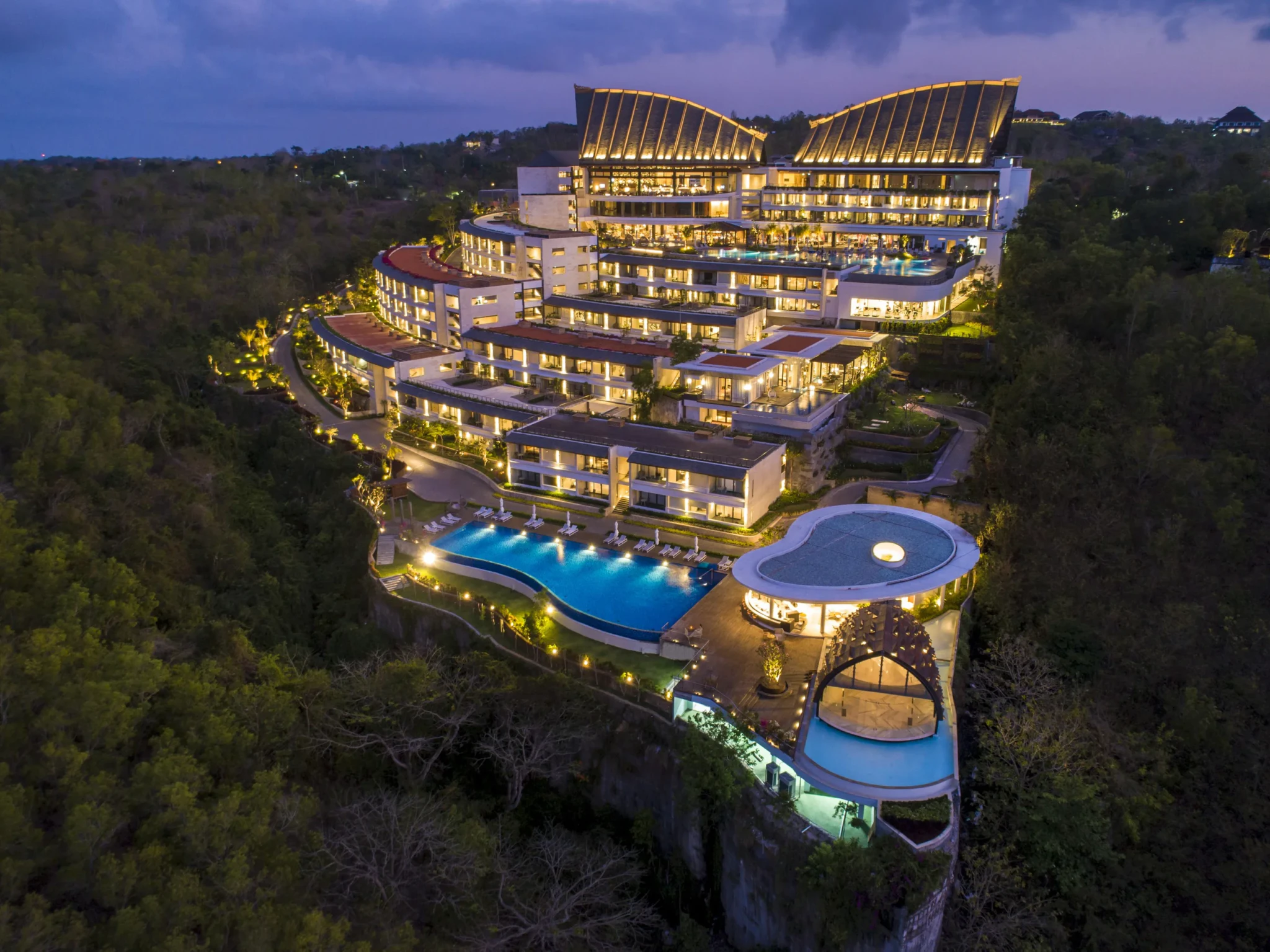 Aerial dusk view of Renaissance Bali Uluwatu Resort & Spa with illuminated curved roofs and a cliff-edge pool above the forest
