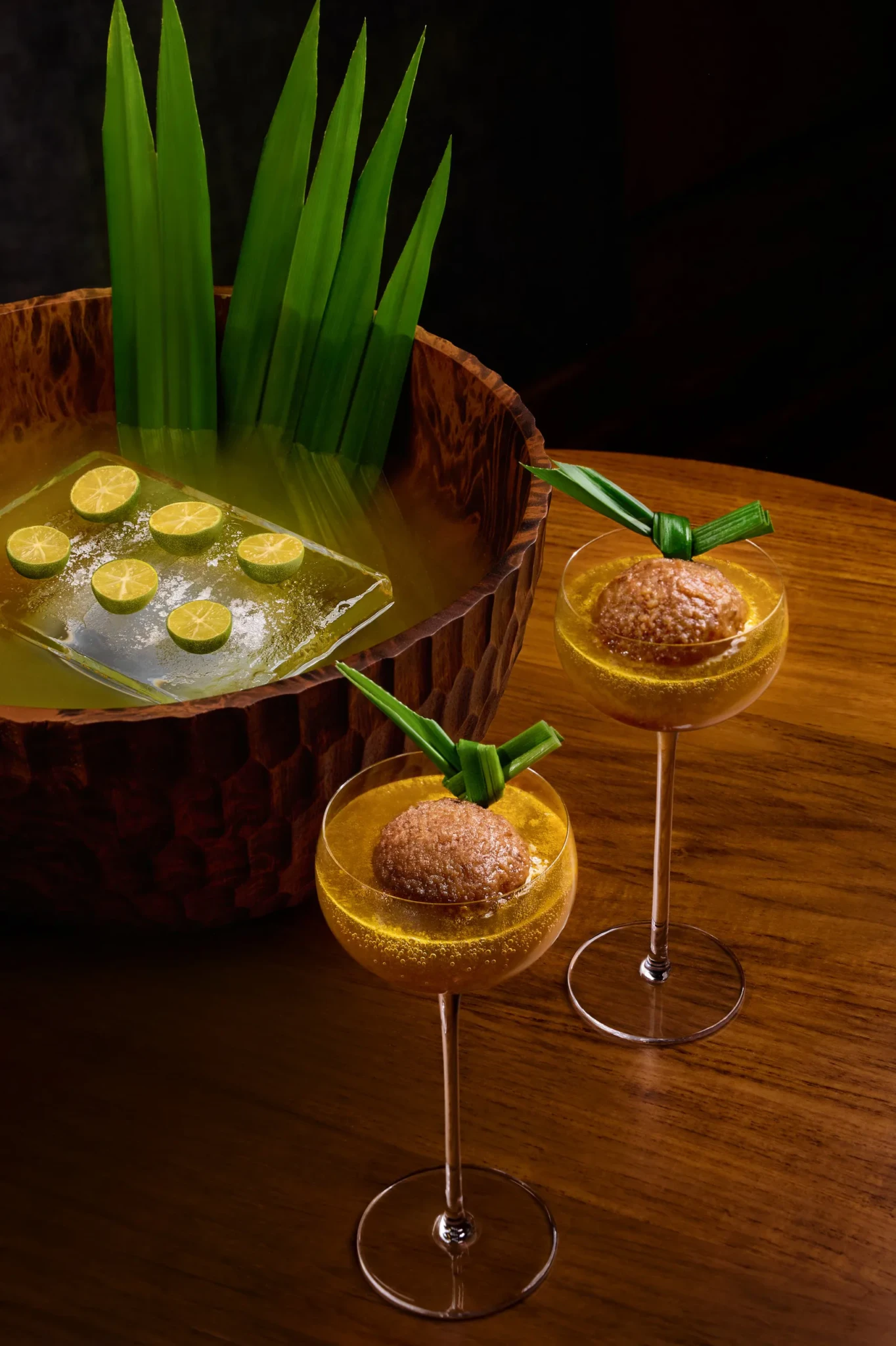 The Night Market punch in gold coupe glasses topped with sugar-dusted spheres and knotted pandan leaves beside a carved wooden punch bowl