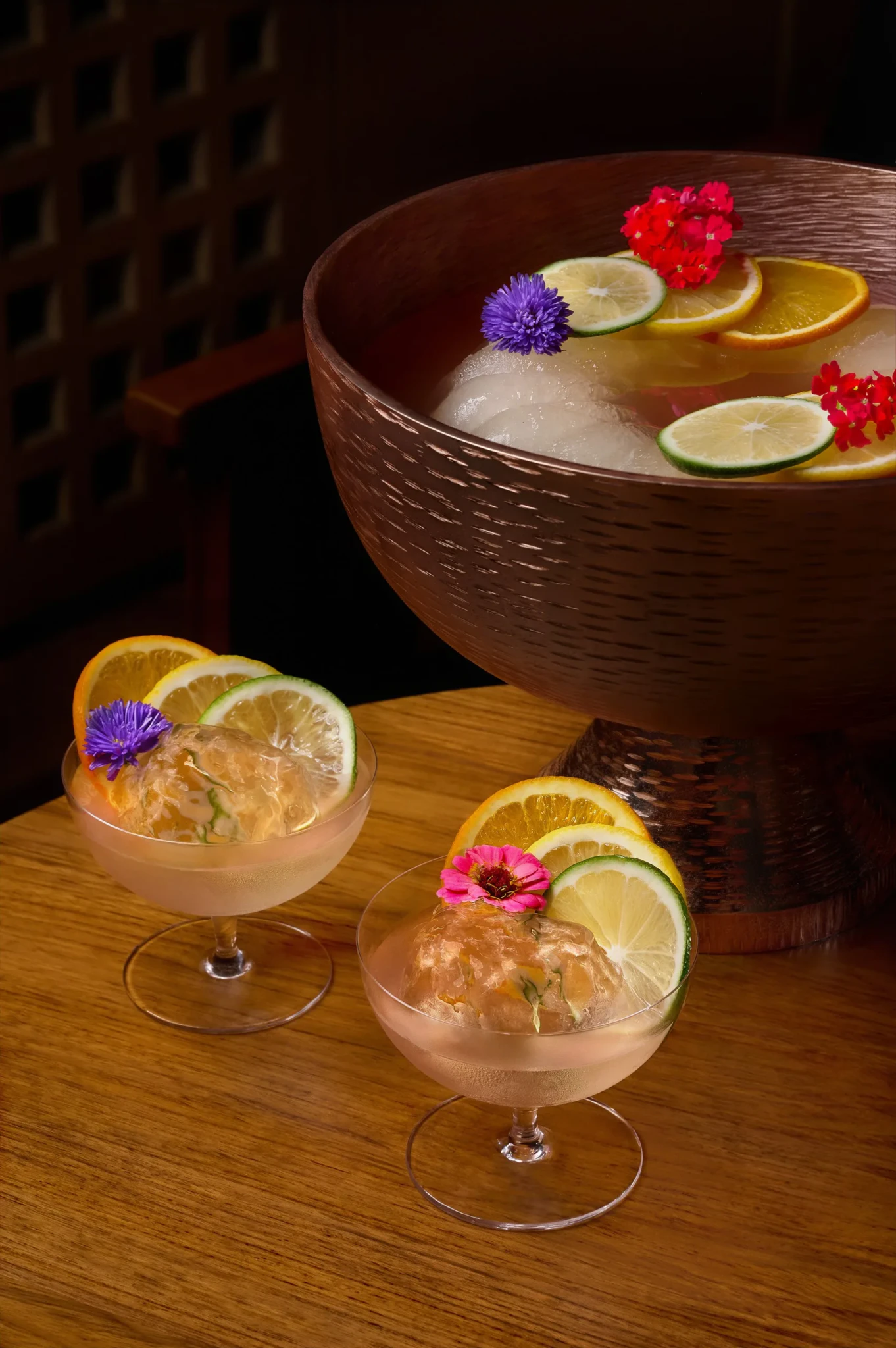 Two coupe glasses of pale pink Blooming Flower punch with ice spheres, citrus slices and edible flowers beside a hammered copper punch bowl