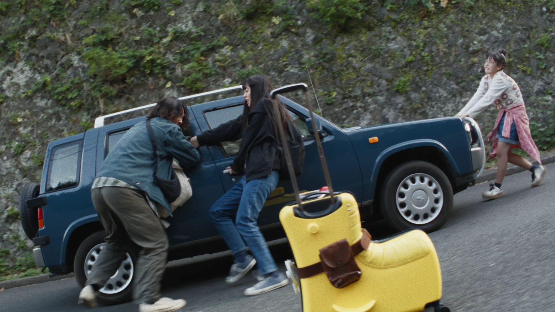 A family loads a dark-blue SUV by the roadside as someone runs towards them, with a yellow suitcase in the foreground