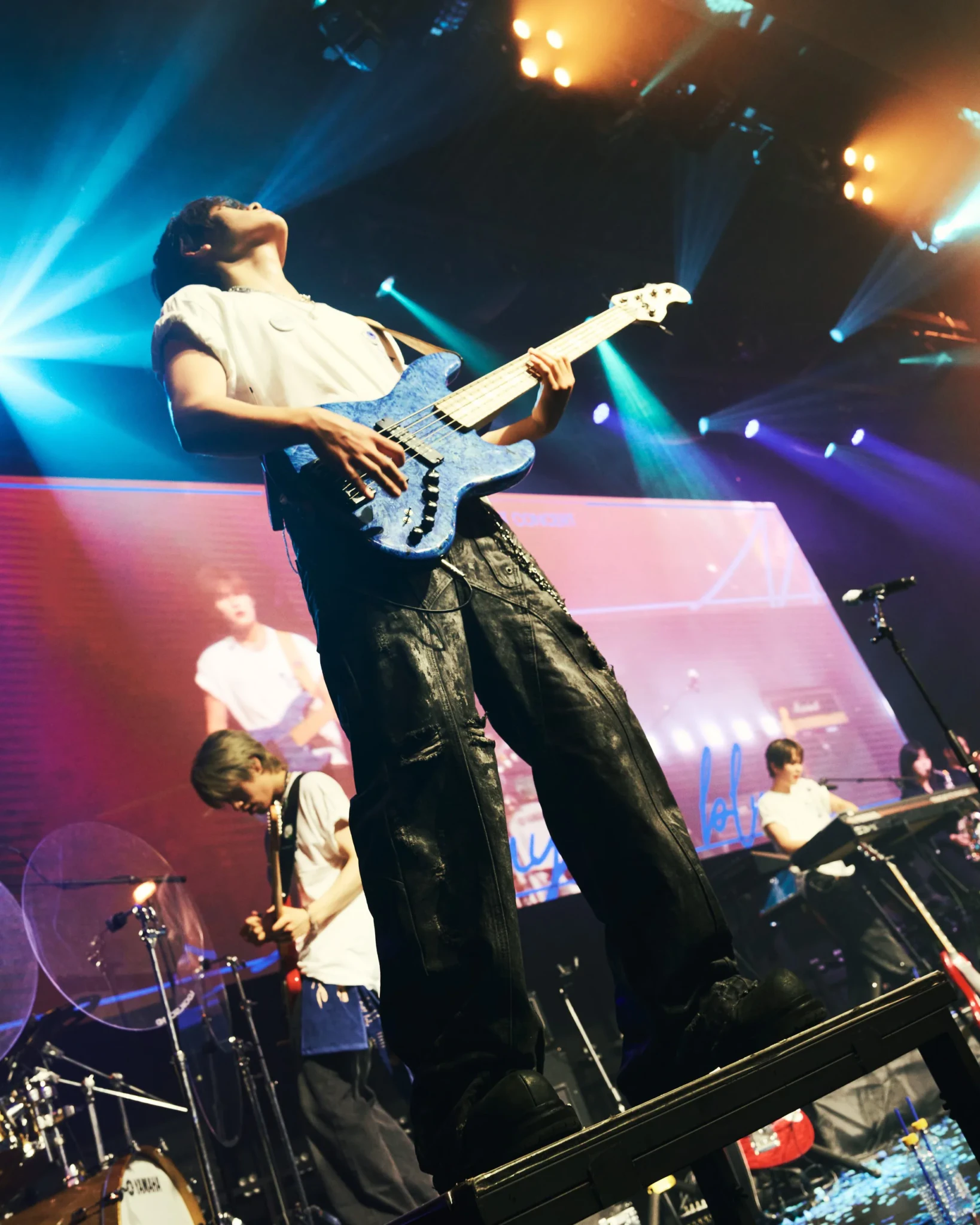 Bassist in white shirt and black pants performing on stage under blue lights, bandmates in background