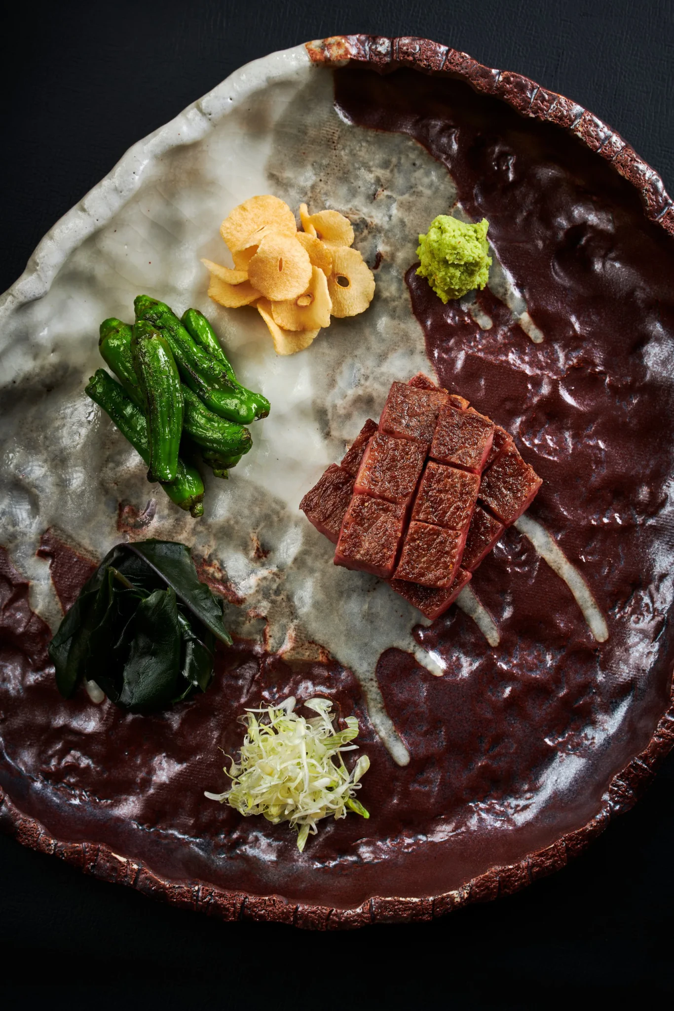 Charcoal-grilled Ohmi Wagyu cut into cubes with shishito peppers, crisp chips, wasabi and pickled greens on a rustic ceramic plate