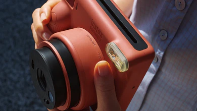 A hand holds the terracotta-coloured instax SQUARE 2 instant camera, showing its lens and built-in flash.