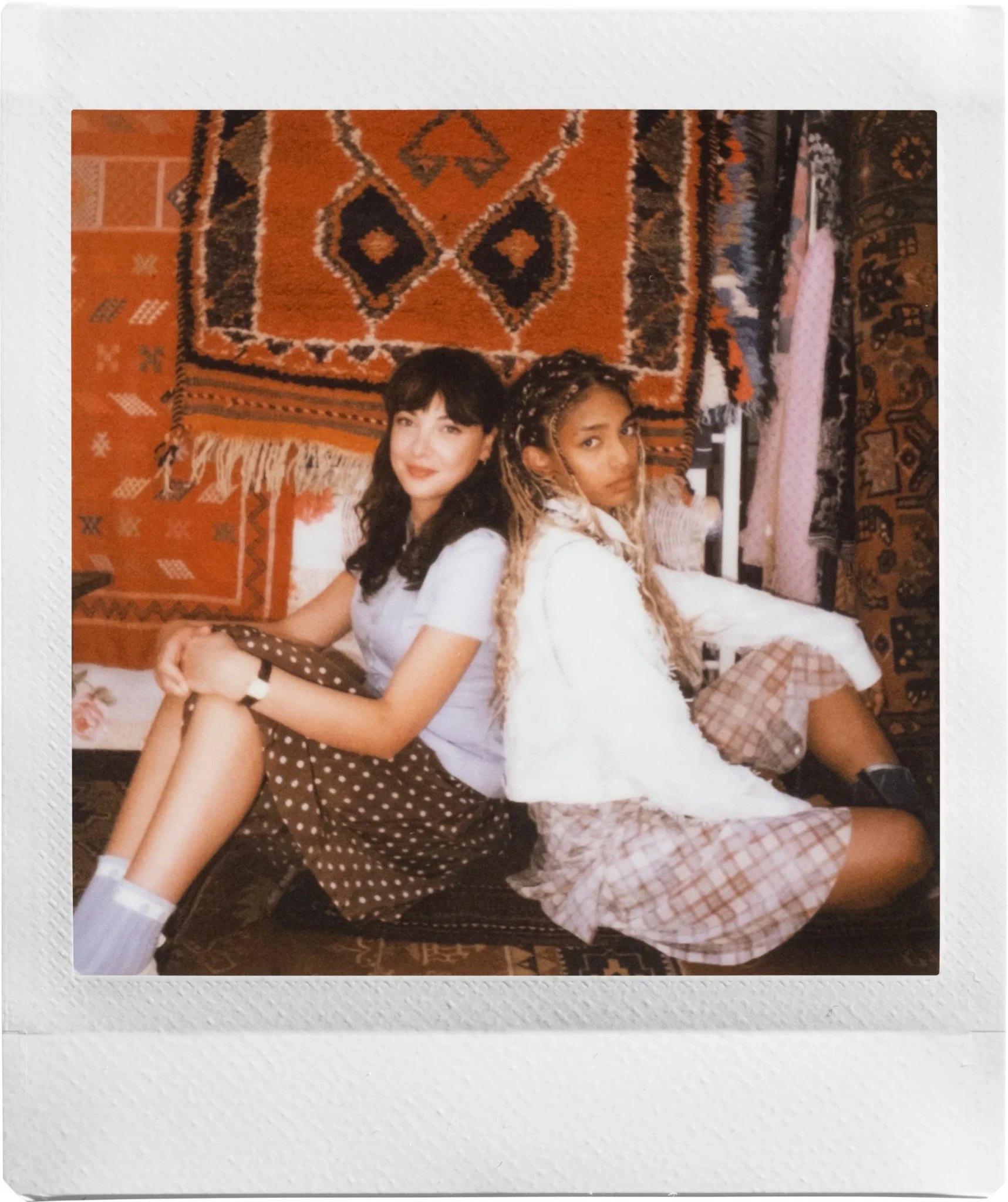 Two young women sitting back to back on a rug in front of a colourful patterned wall hanging, an instax SQUARE 2 sample print