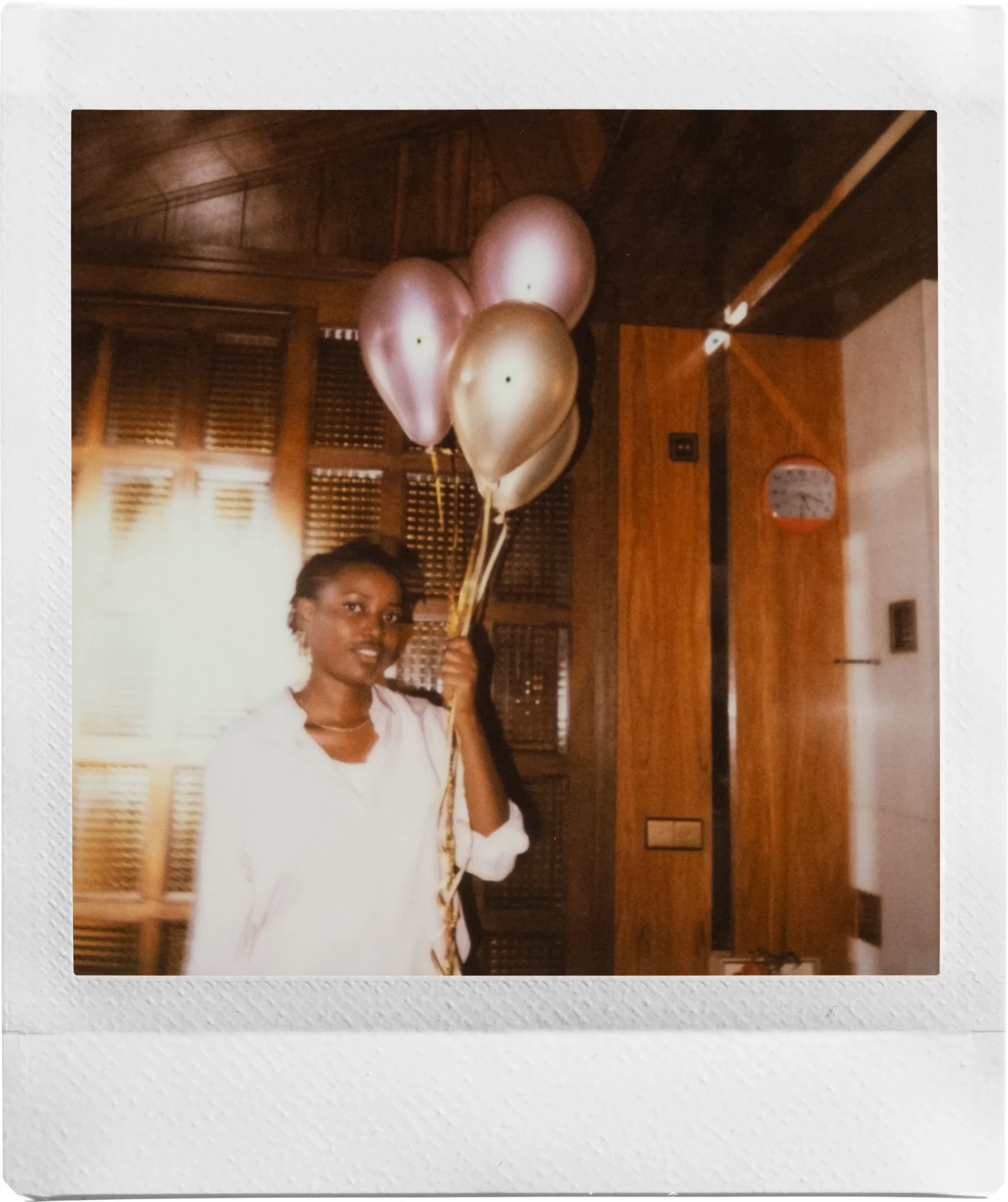 A woman in a white shirt holding pink and gold balloons in a warmly lit room with wood-panelled walls