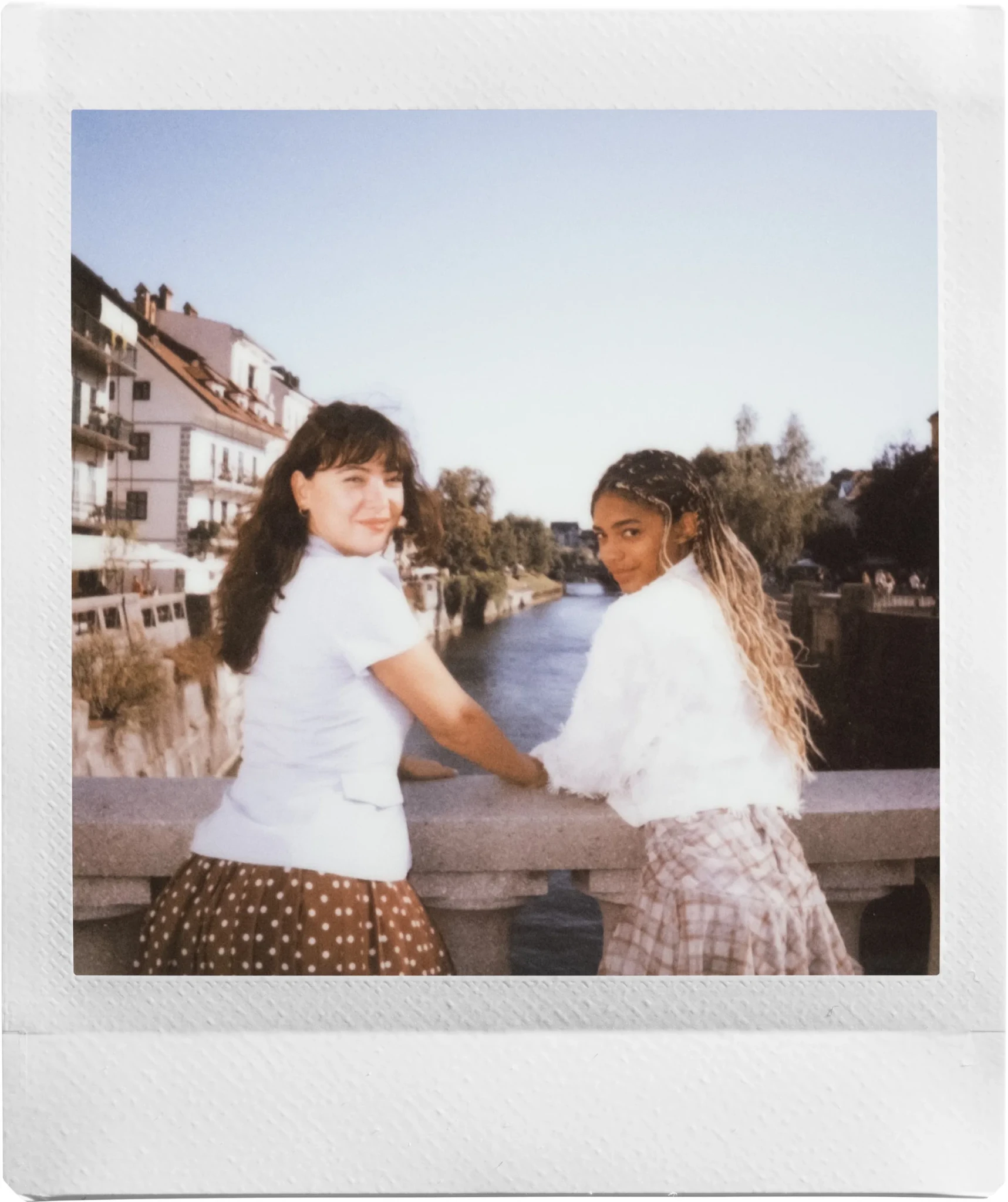 Two women with their arms linked on a stone railing beside a river, an instax SQUARE sample print