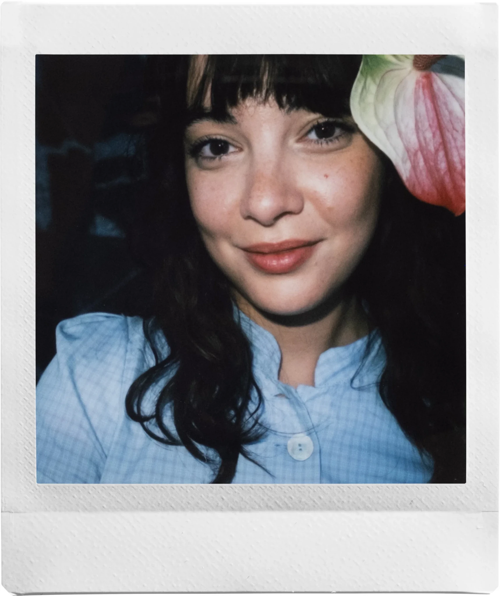 A smiling woman with dark bangs and long wavy hair next to a pink-and-white anthurium flower in a square instant print