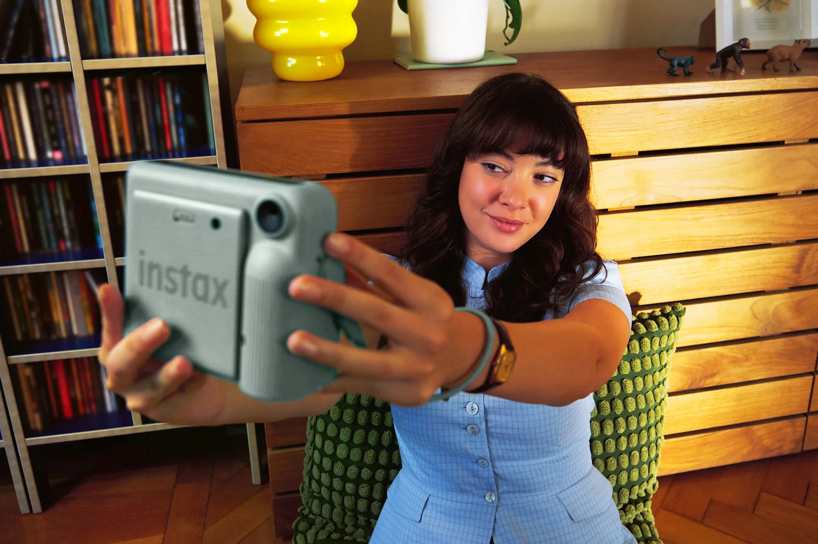 A woman with dark bangs takes a selfie with a mint-green instax camera in a warmly lit room with bookshelves and a wooden dresser.