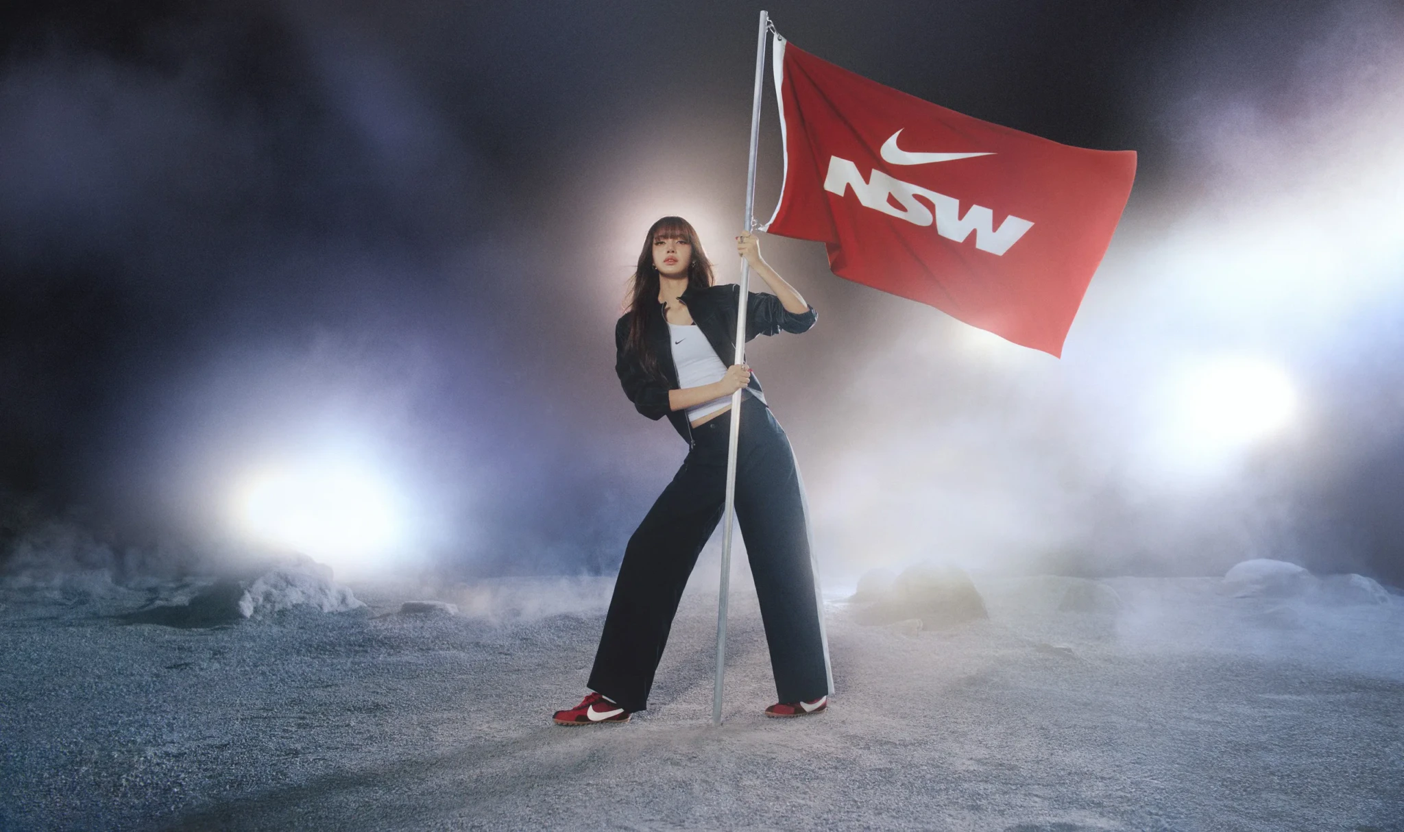 Woman in black track pants and white crop top holding a red Nike NSW flag in a foggy, moonlike landscape