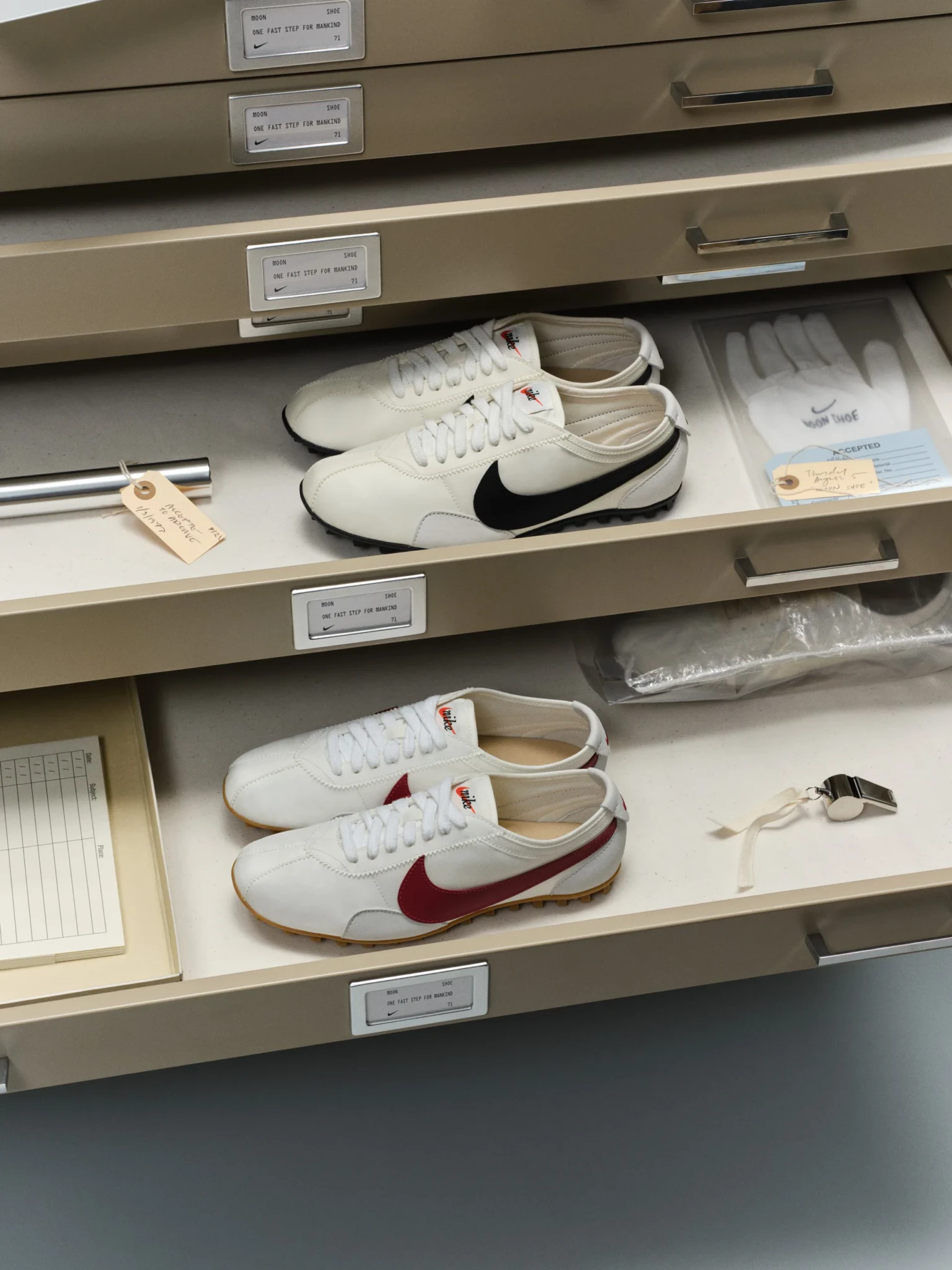 Two pairs of Nike Moon Shoes in black and white, and white and red, in an archival drawer with a glove and whistle
