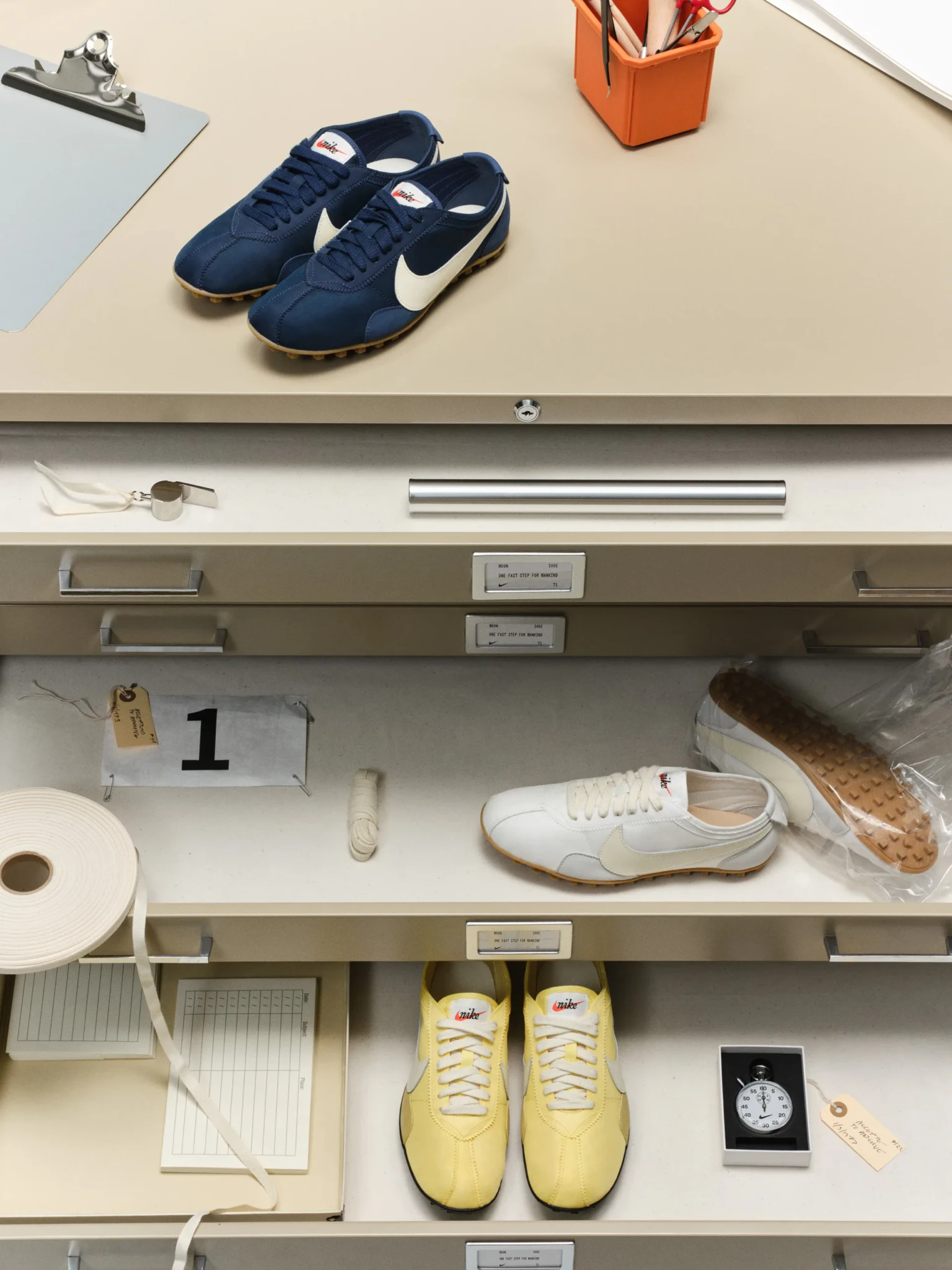 Nike Moon Shoe sneakers in navy, white and yellow displayed on archive-style drawers with props including a stopwatch and tags.