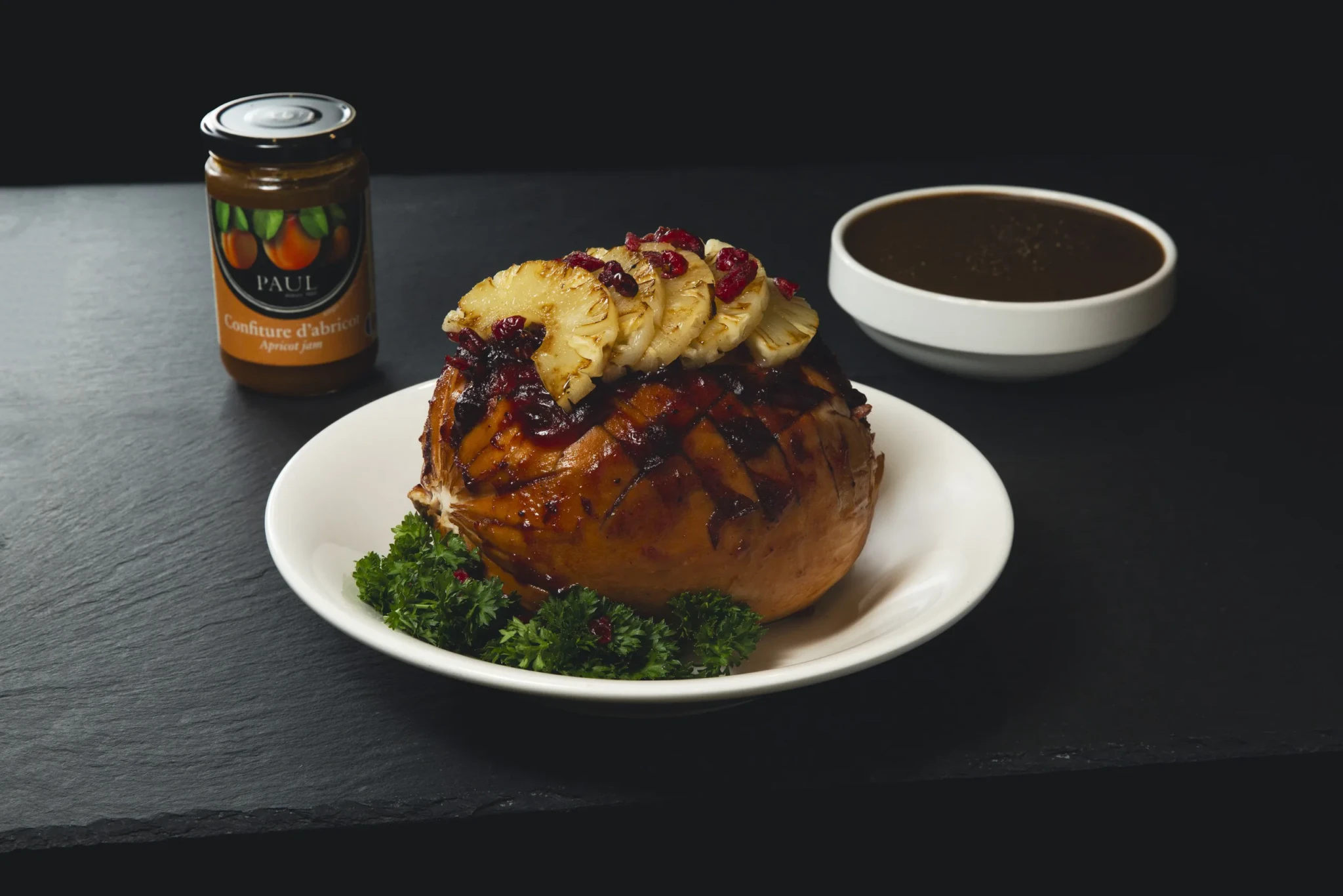 Glazed cranberry and pineapple turkey ham on a white plate with parsley, beside a jar of PAUL apricot jam and a bowl of gravy.