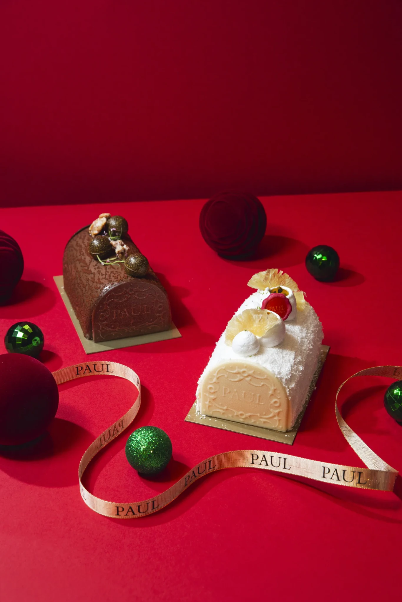 Two PAUL Yule logs, a walnut chocolate log and a white tropical fruit log, on red background with festive baubles and PAUL ribbon