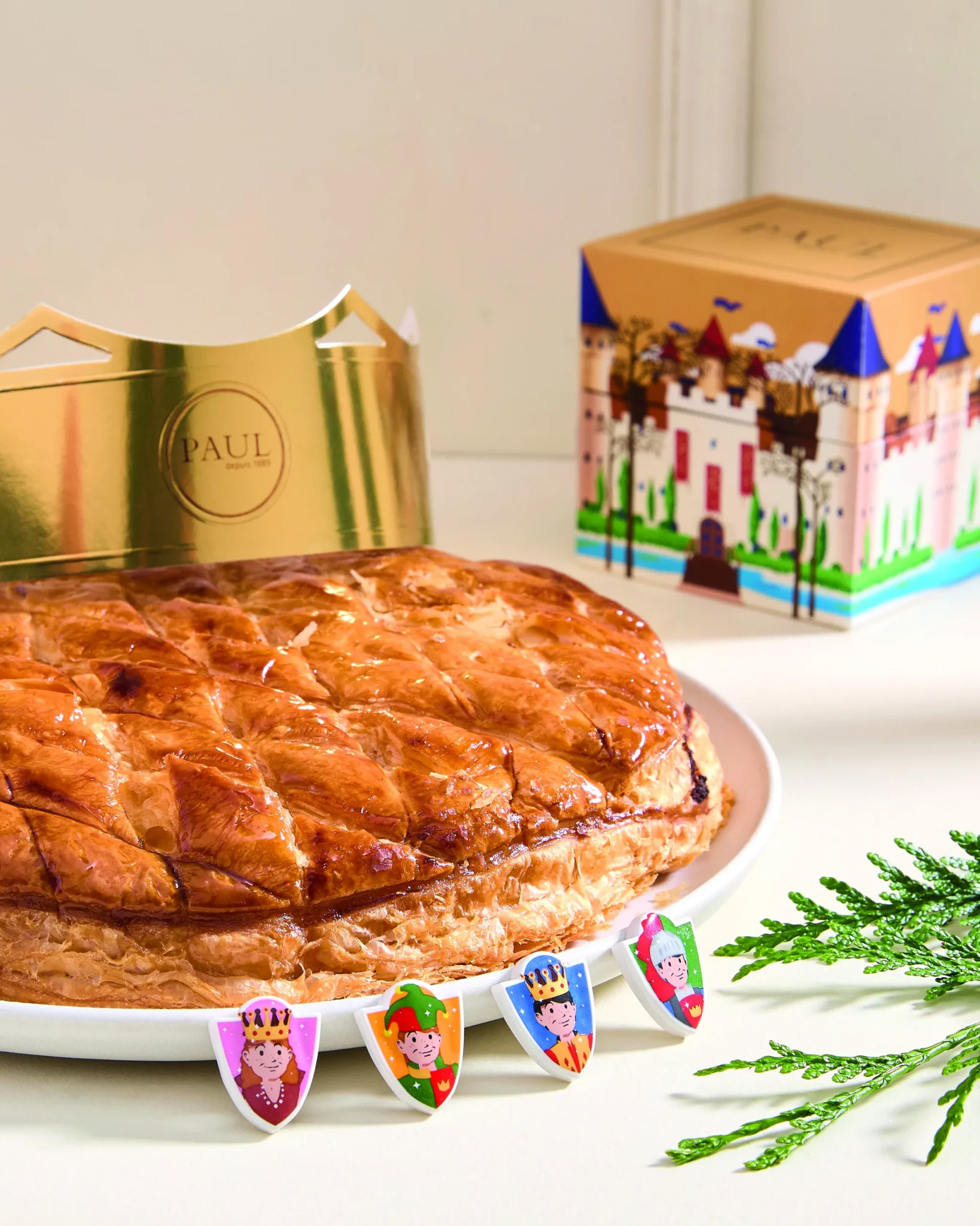 A golden Galette des Rois with colourful character figurines on the plate and a crown-themed PAUL box behind it