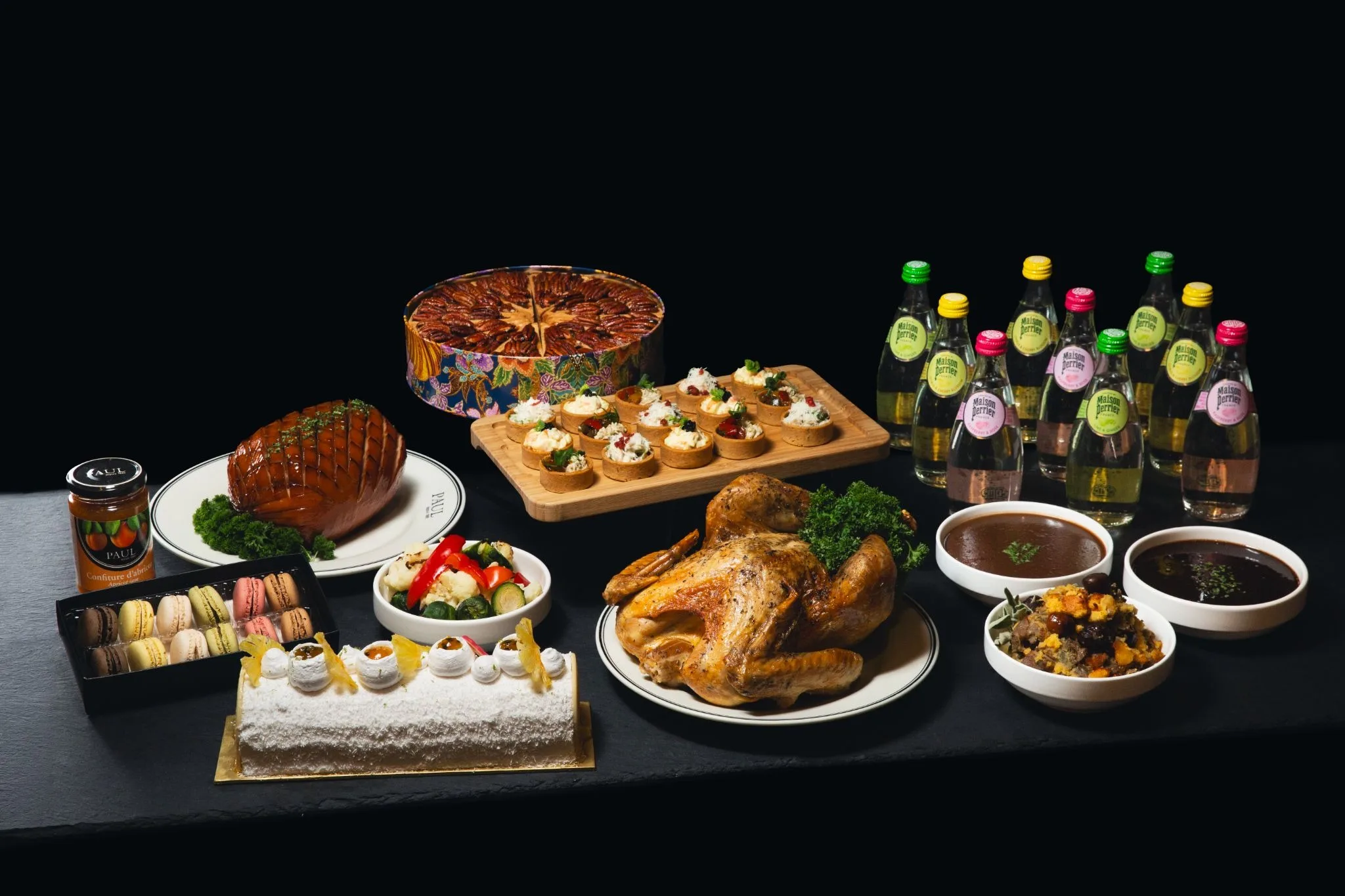 PAUL's Noël Collection 2026 spread featuring a roast turkey, glazed ham, tartlets, a pecan tart, a Yule log, macarons, and Maison Perrier sparkling water.