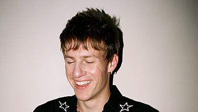 Malcolm Todd smiling with his eyes closed against a plain wall, wearing a black shirt with white star embroidery