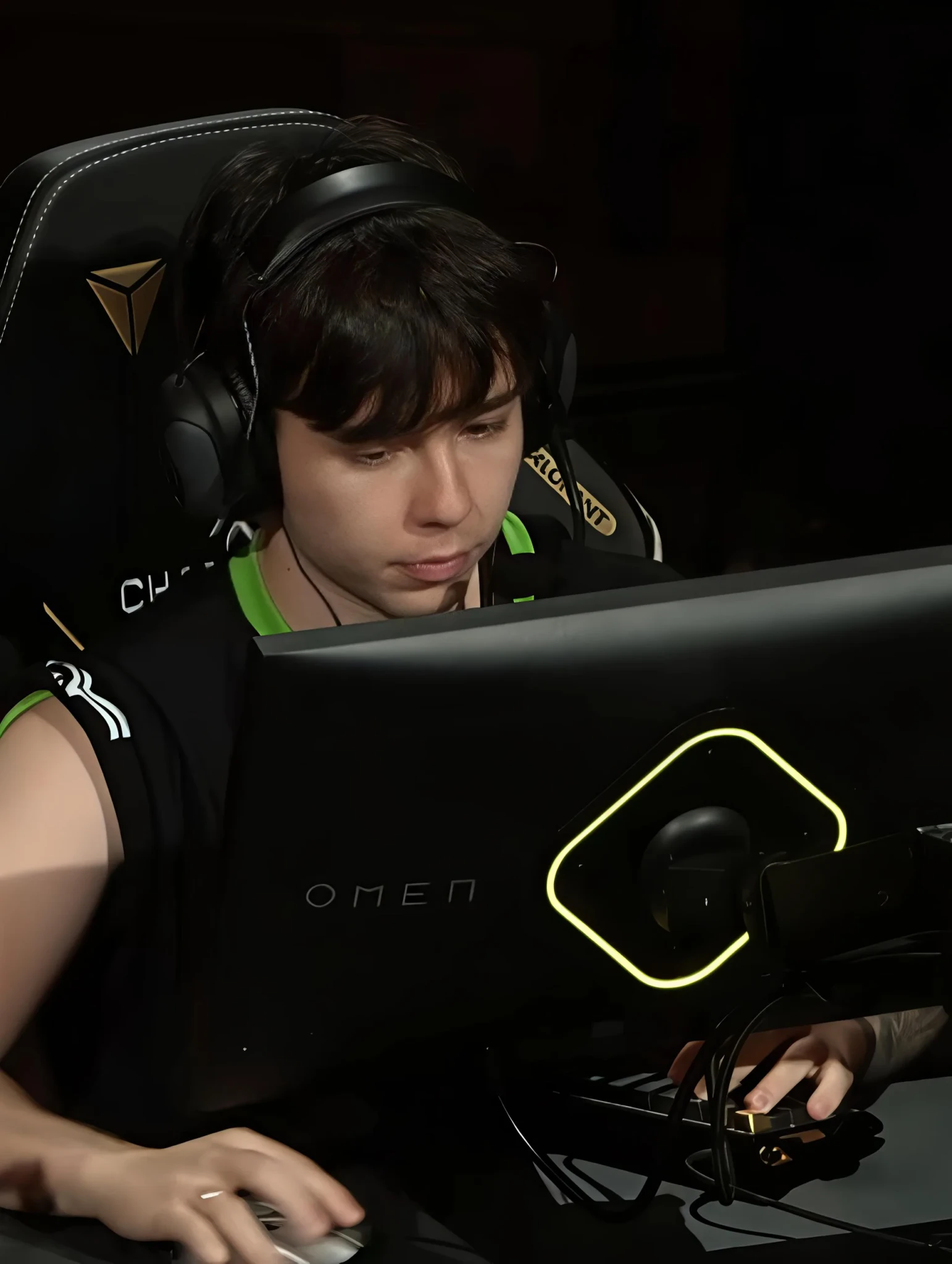 Gamer in headphones and black-green jersey focused on an OMEN monitor, with a yellow-lit logo and keyboard in view