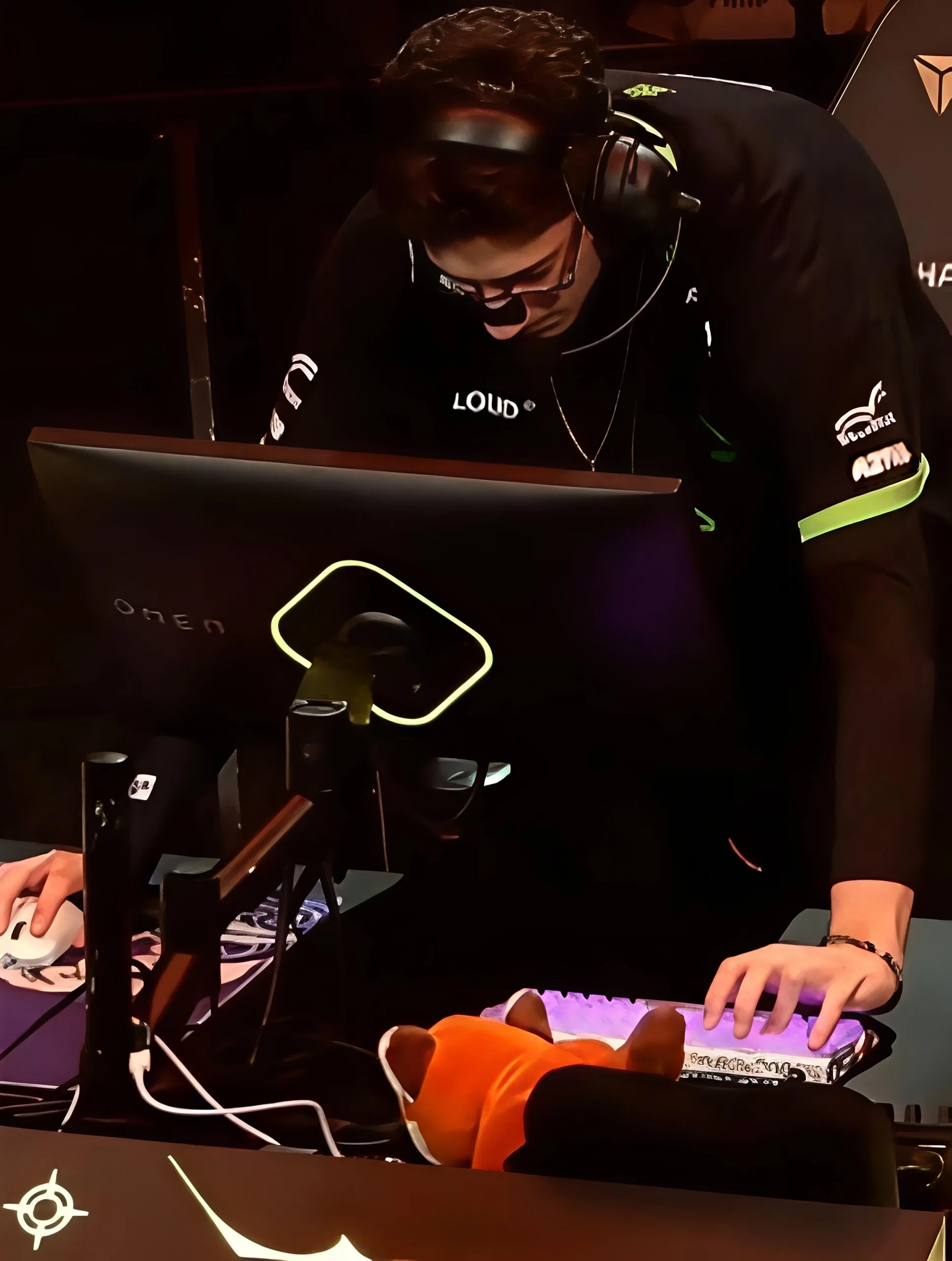 A LOUD player leans toward his monitor mid-match, an orange plush toy beside a lit keyboard on the desk