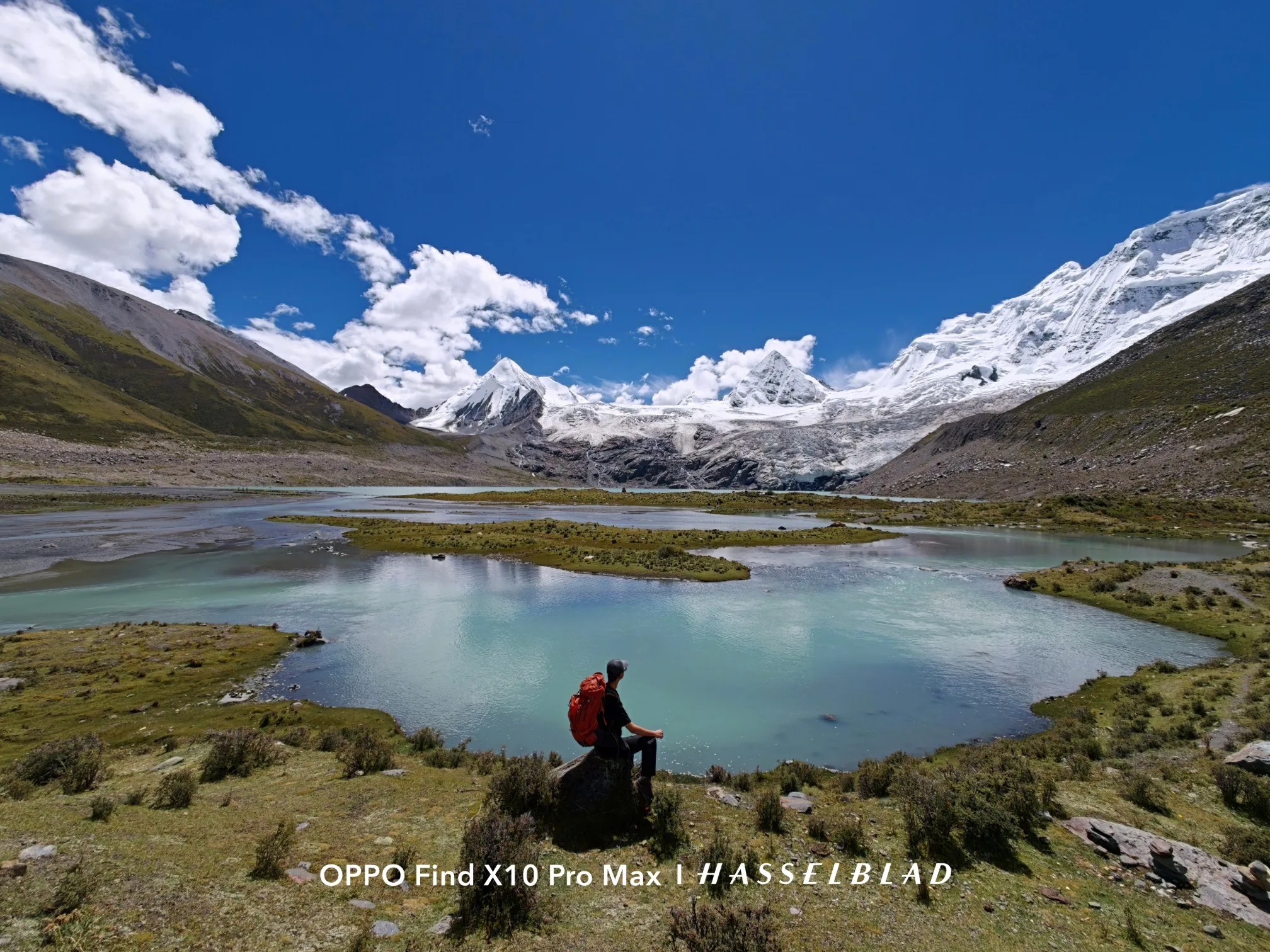 A hiker with a red backpack sitting above a turquoise glacial lake beneath snowy peaks, shot on the Find X10 Pro Max