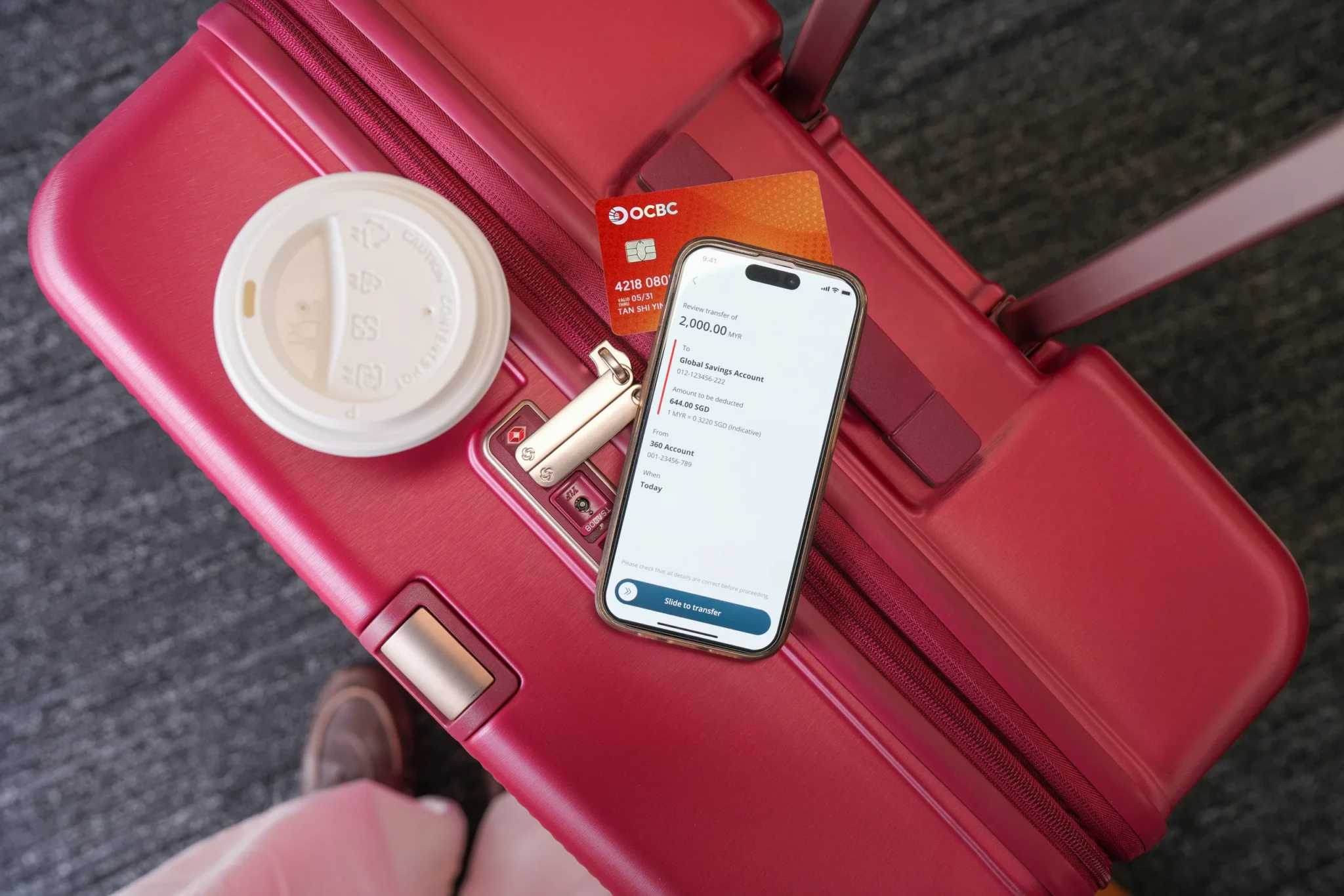 A red OCBC Global Savings Account debit card and a phone showing a MYR 2,000 transfer review screen rest on a red suitcase beside a coffee cup
