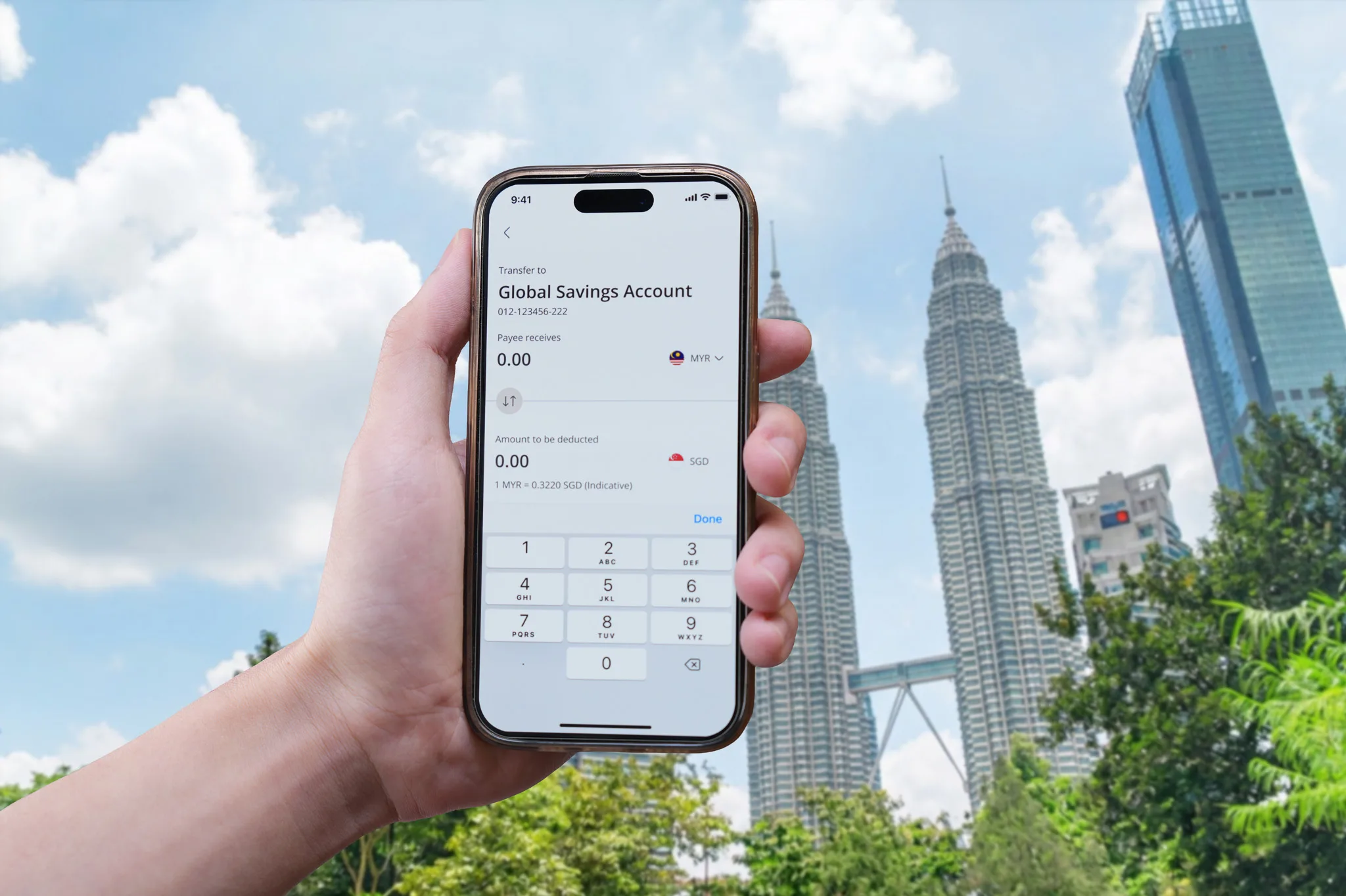 A hand holds a phone showing the OCBC app's transfer screen for a Global Savings Account in front of the Petronas Twin Towers