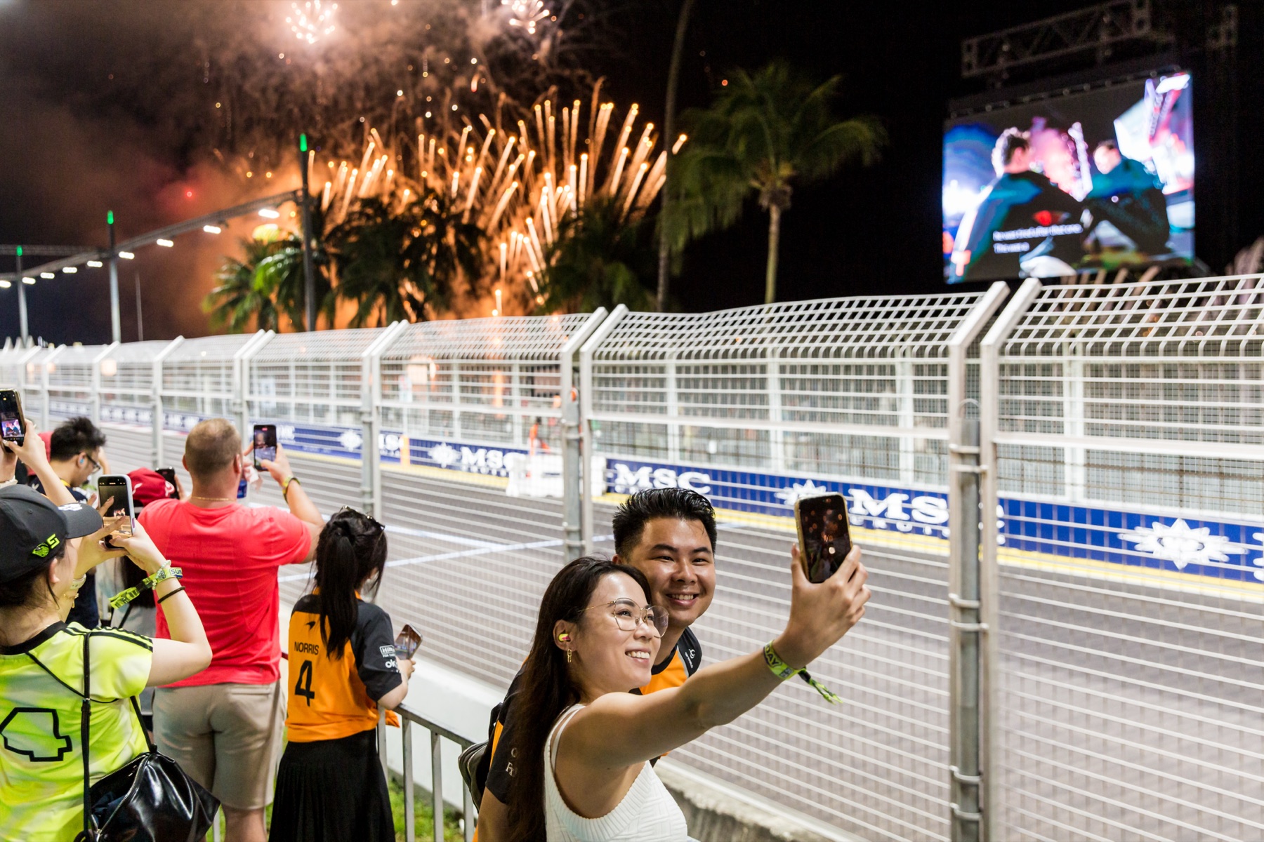Fans take selfies at the circuit fence in front of the Zone 1 Skyline Grandstand, with fireworks above the Marina Bay Street Circuit.