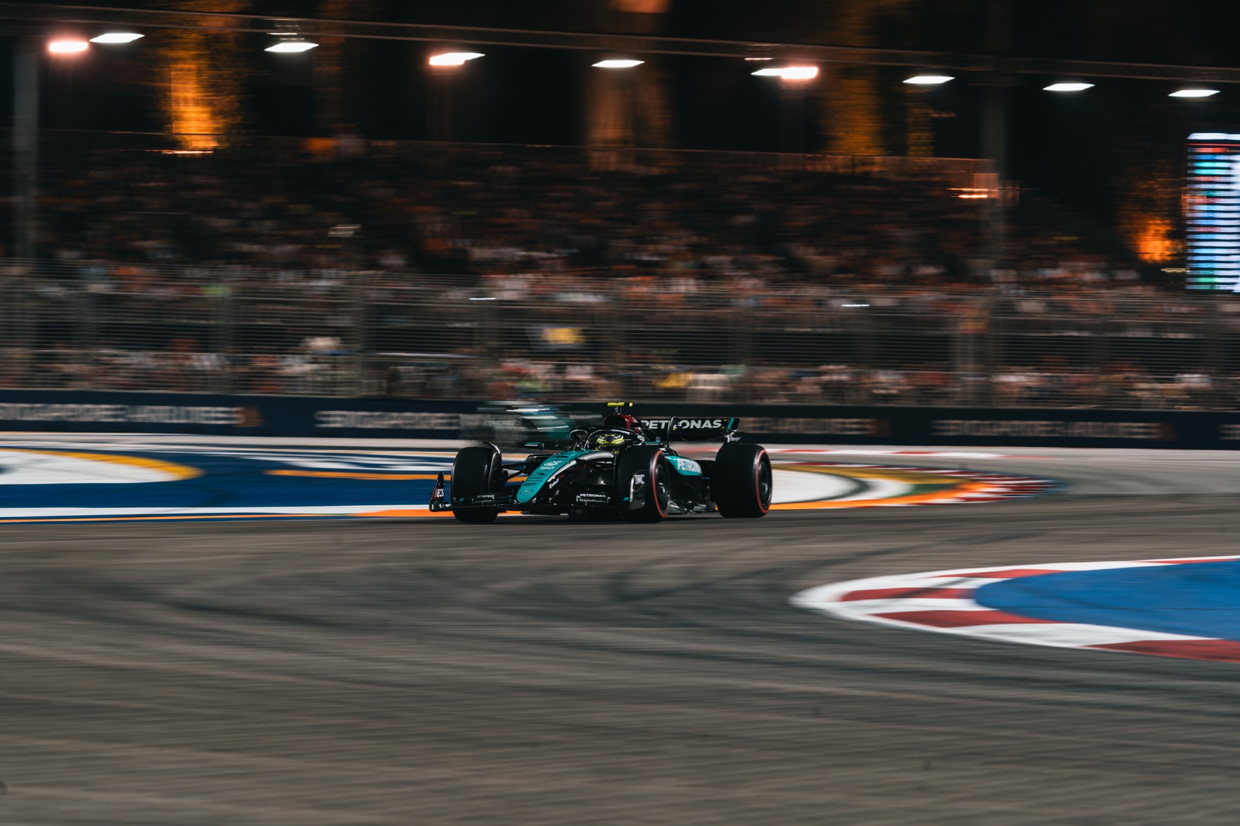 A Mercedes Formula 1 car cornering at night at the Marina Bay Street Circuit, with floodlit spectators behind.