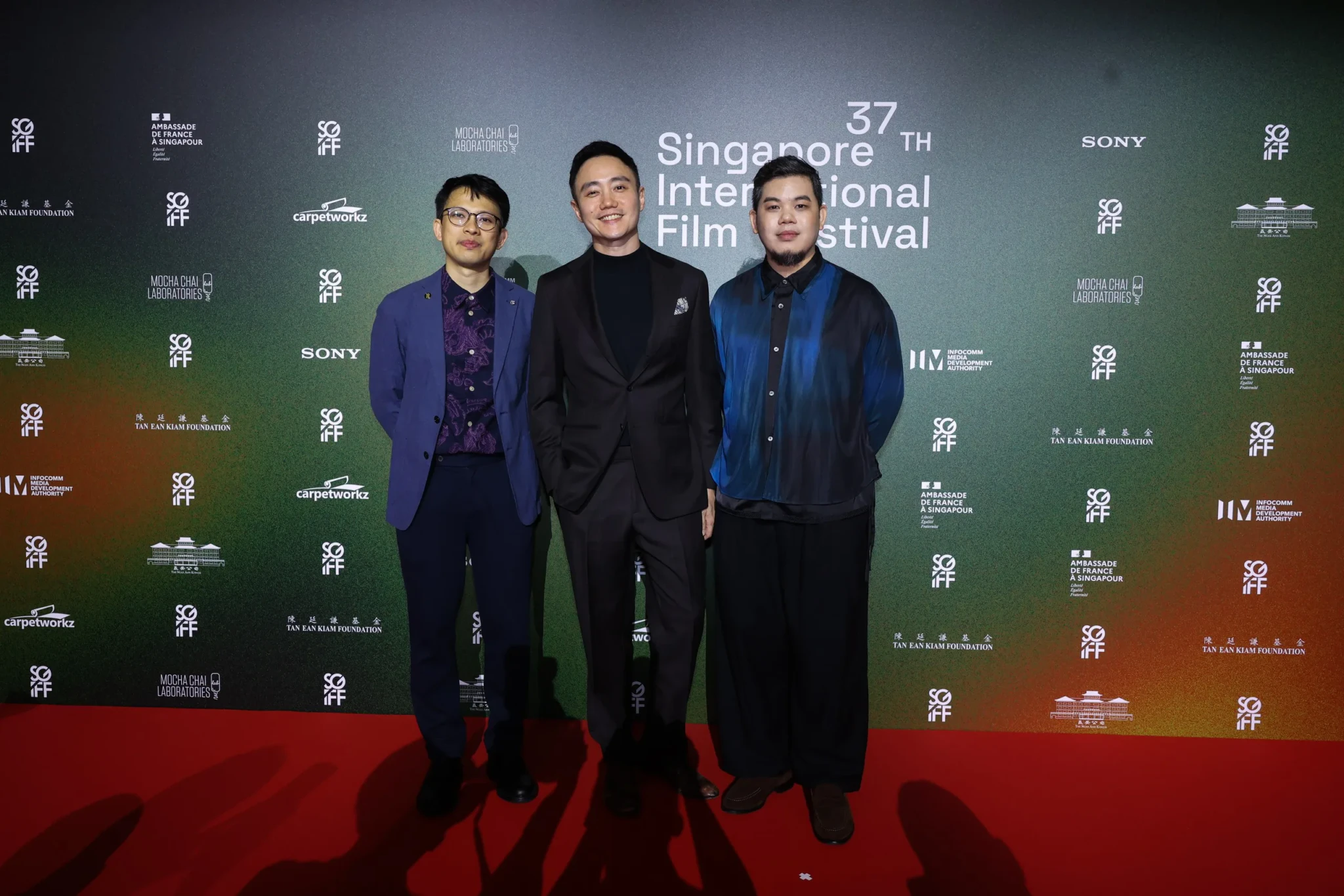 Three guests pose together on the red carpet in front of the 37th Singapore International Film Festival backdrop