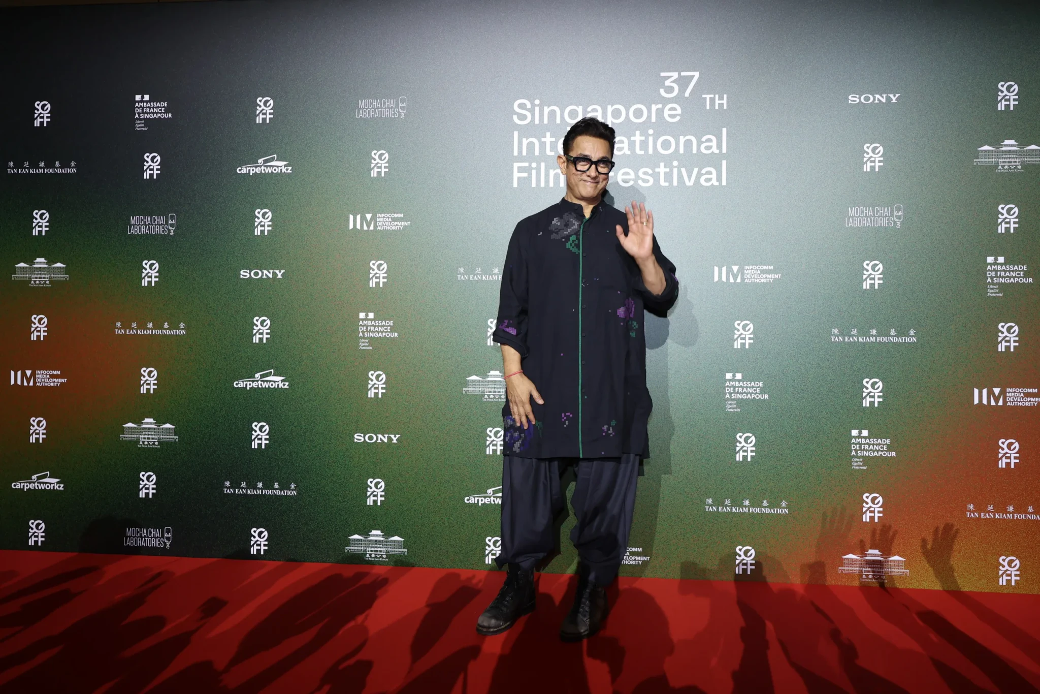 A man in a dark embroidered shirt and glasses waves on the red carpet at the 37th Singapore International Film Festival