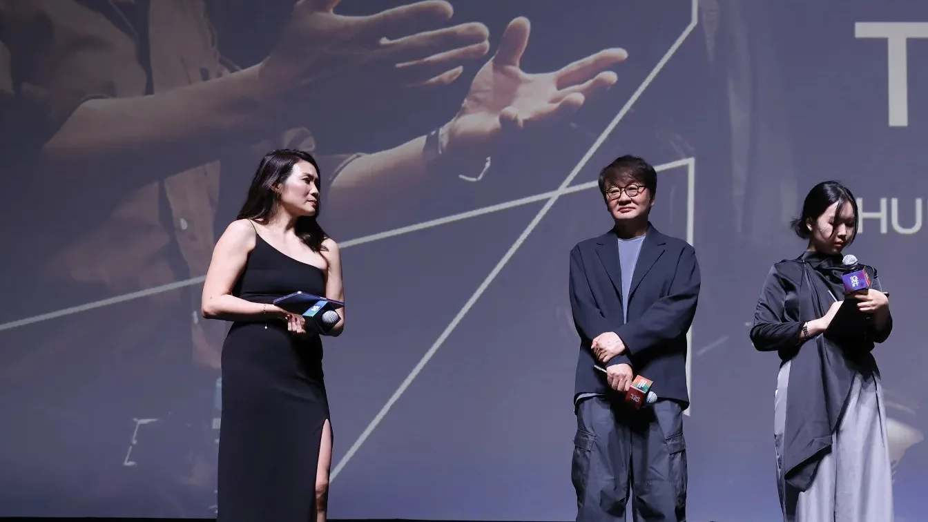 Three people on a festival stage in front of a large projected image of hands gesturing, with a partial festival title visible.