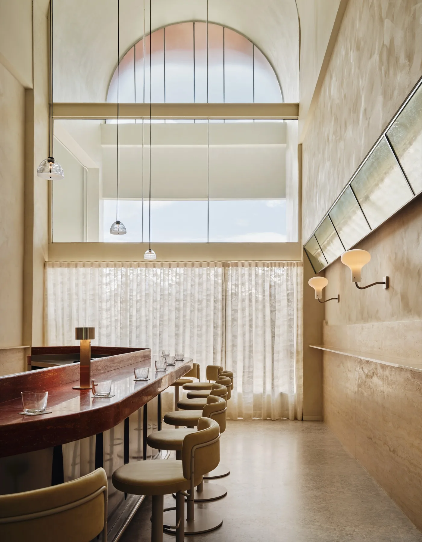 The curved red marble bar counter at Bar Somma with beige velvet stools and a tall arched window