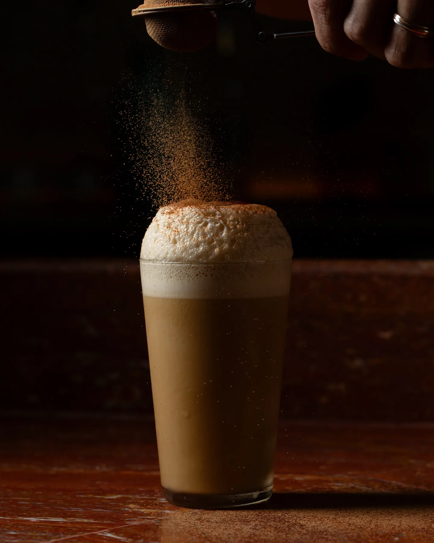 A frothy cocktail in a tall glass being dusted with powder through a fine mesh strainer