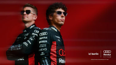 Two Audi Revolut F1 Team drivers in race suits and sunglasses stand back to back against a red background