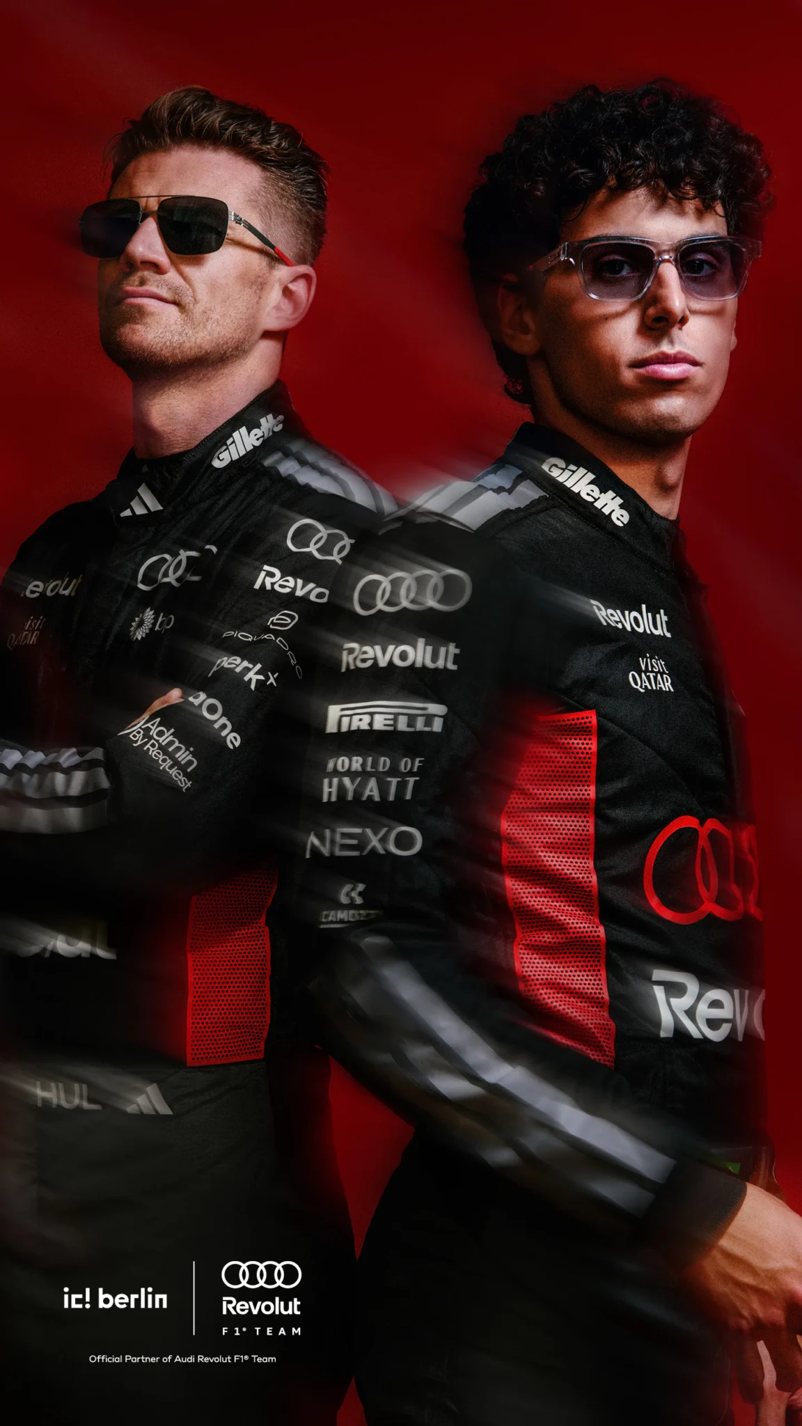 Two men in black Audi Revolut F1 race suits with sponsor logos stand back to back against a red background, one wearing aviator sunglasses.