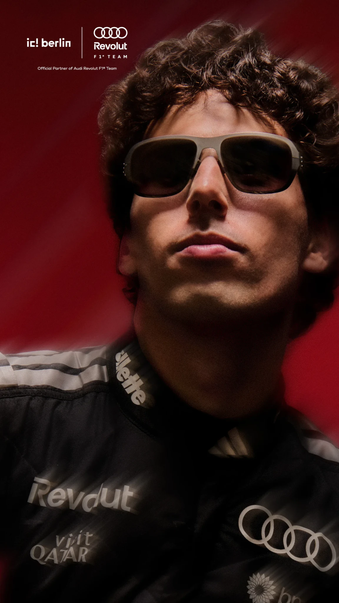 A driver in a black Audi Revolut F1 Team jacket wears tan-framed sunglasses against a red-lit background