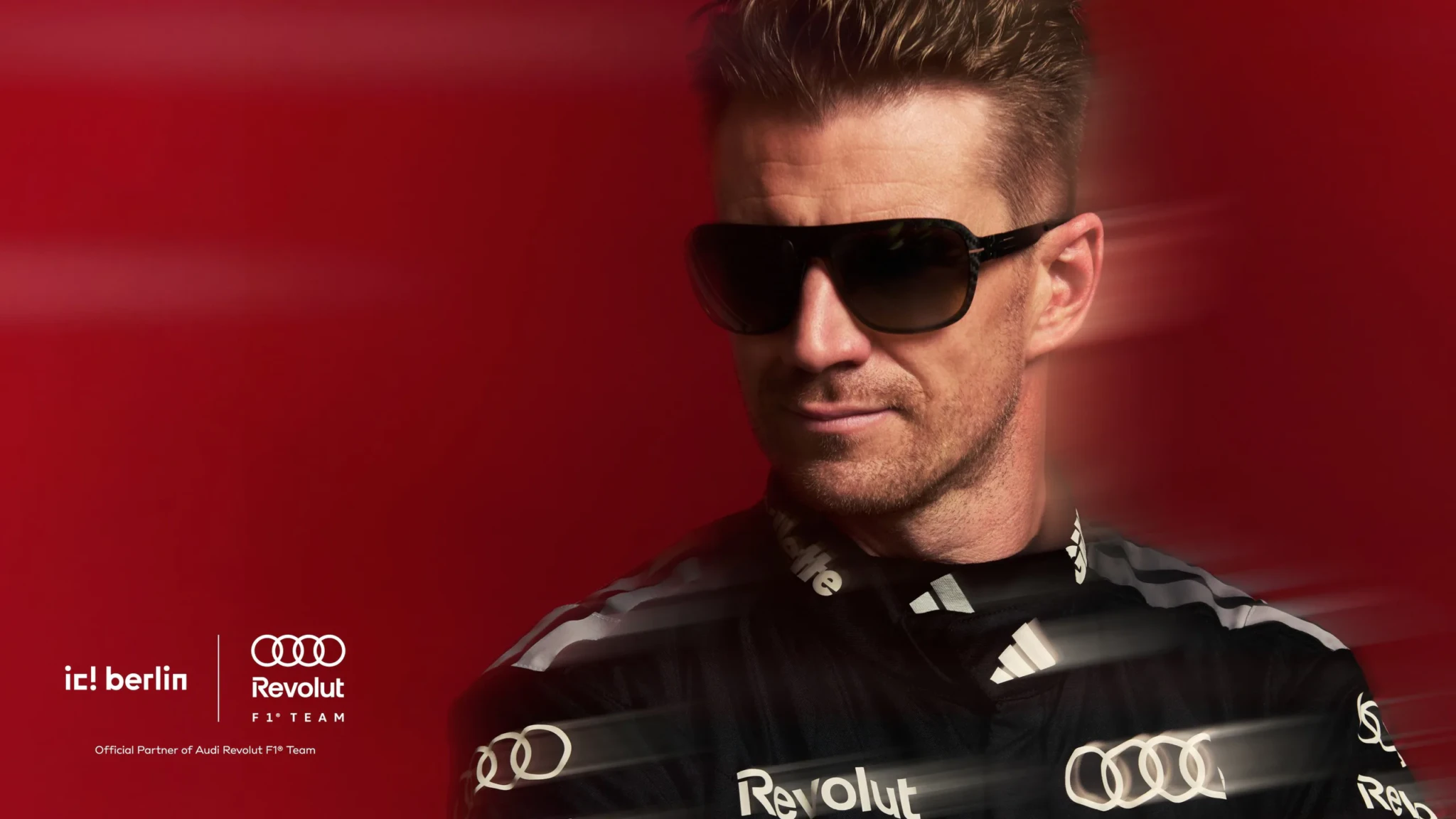 Man in tinted sunglasses and black Audi Revolut F1 Team jacket against a red background with ic! berlin branding