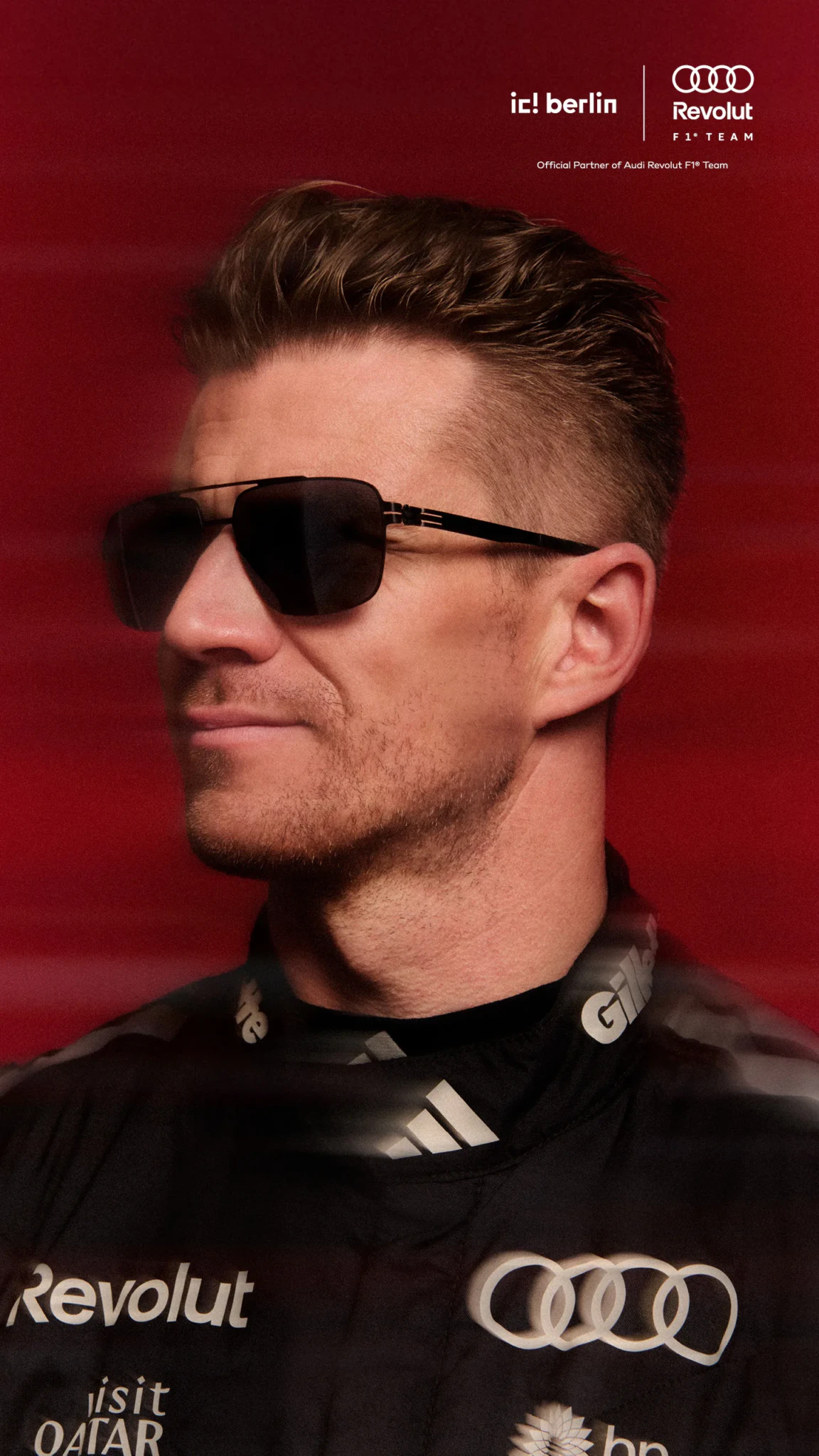 A man in dark aviator-style sunglasses and a black Audi Revolut F1 Team jacket poses against a red background