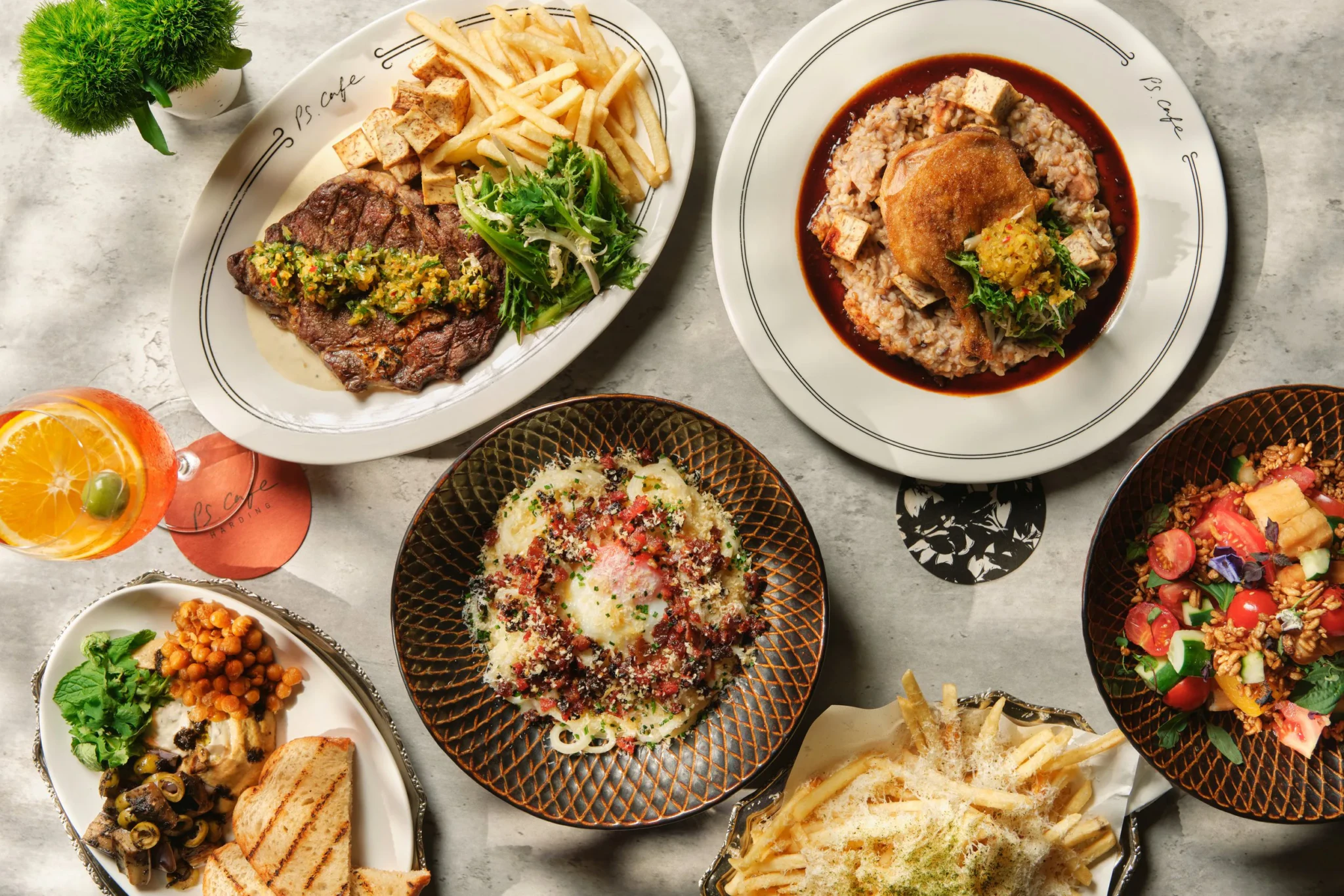 Overhead view of PS.Cafe Harding dishes on a grey table: steak, duck confit risotto, carbonara, salad, fries and hummus.