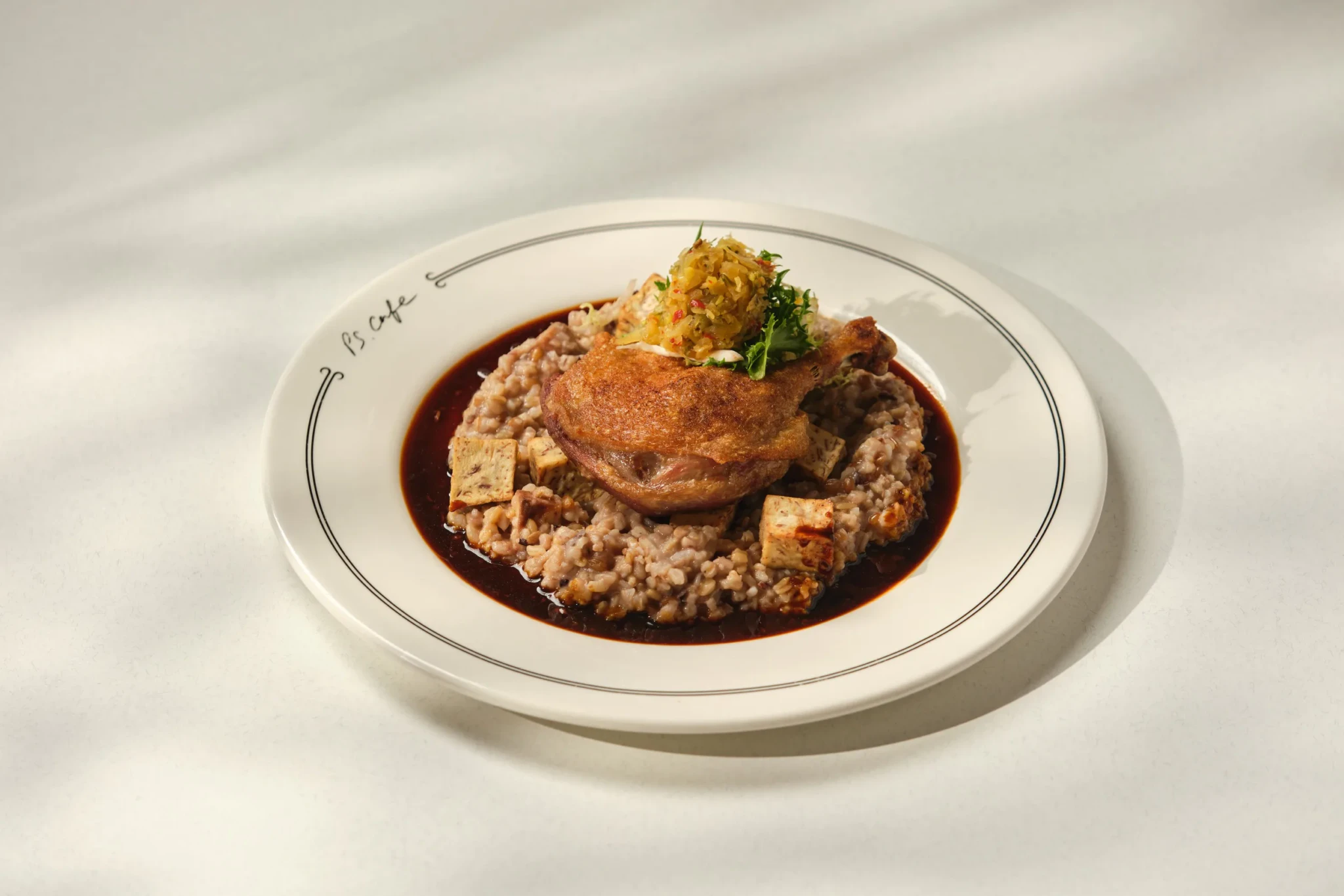Crispy duck confit leg on a bed of risotto with dark jus, cubes of tofu and pickled relish, served on a PS.Cafe plate