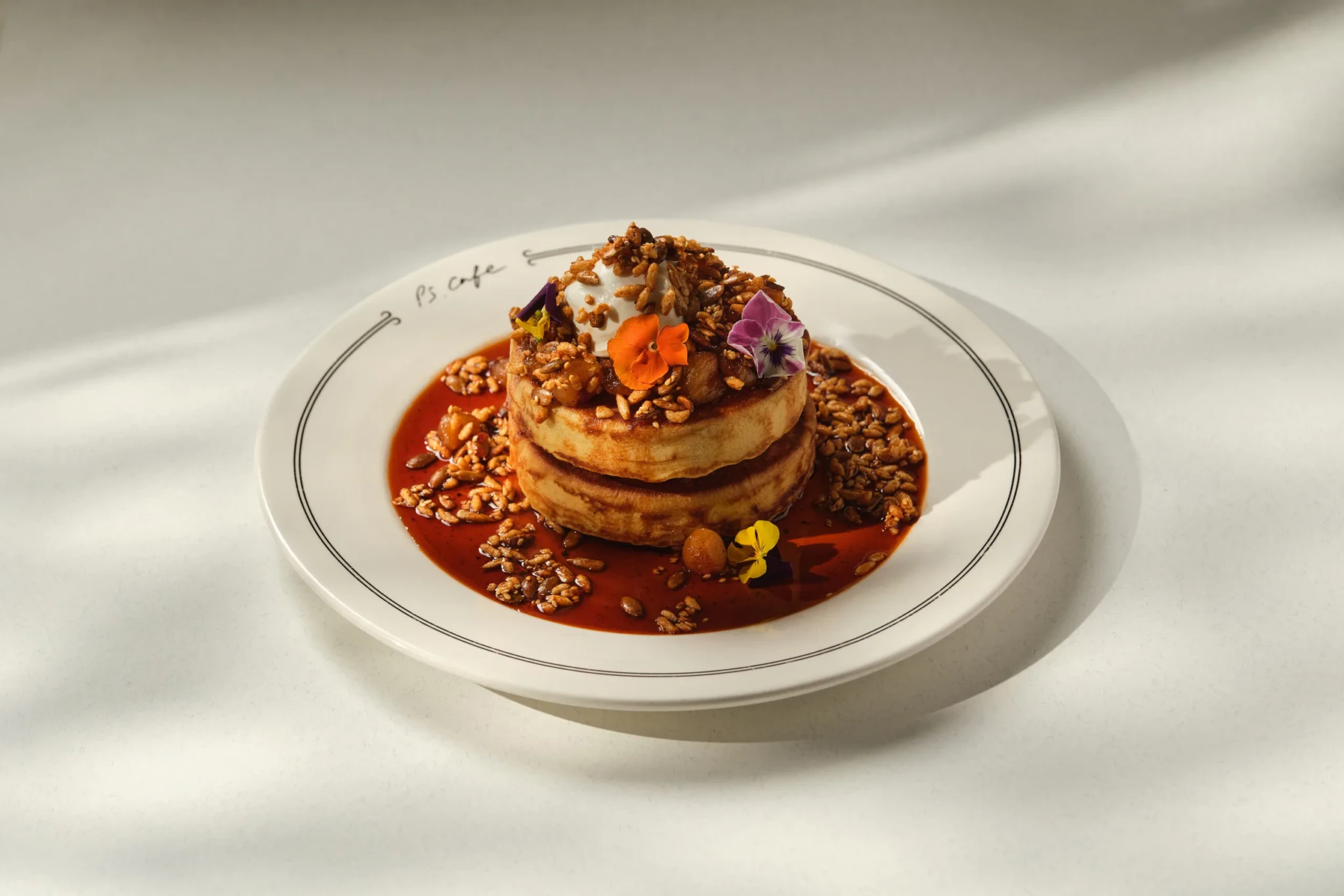 Stacked pancakes with cream, crunchy puffed grain topping, edible flowers and dark caramel sauce on a PS.Cafe plate