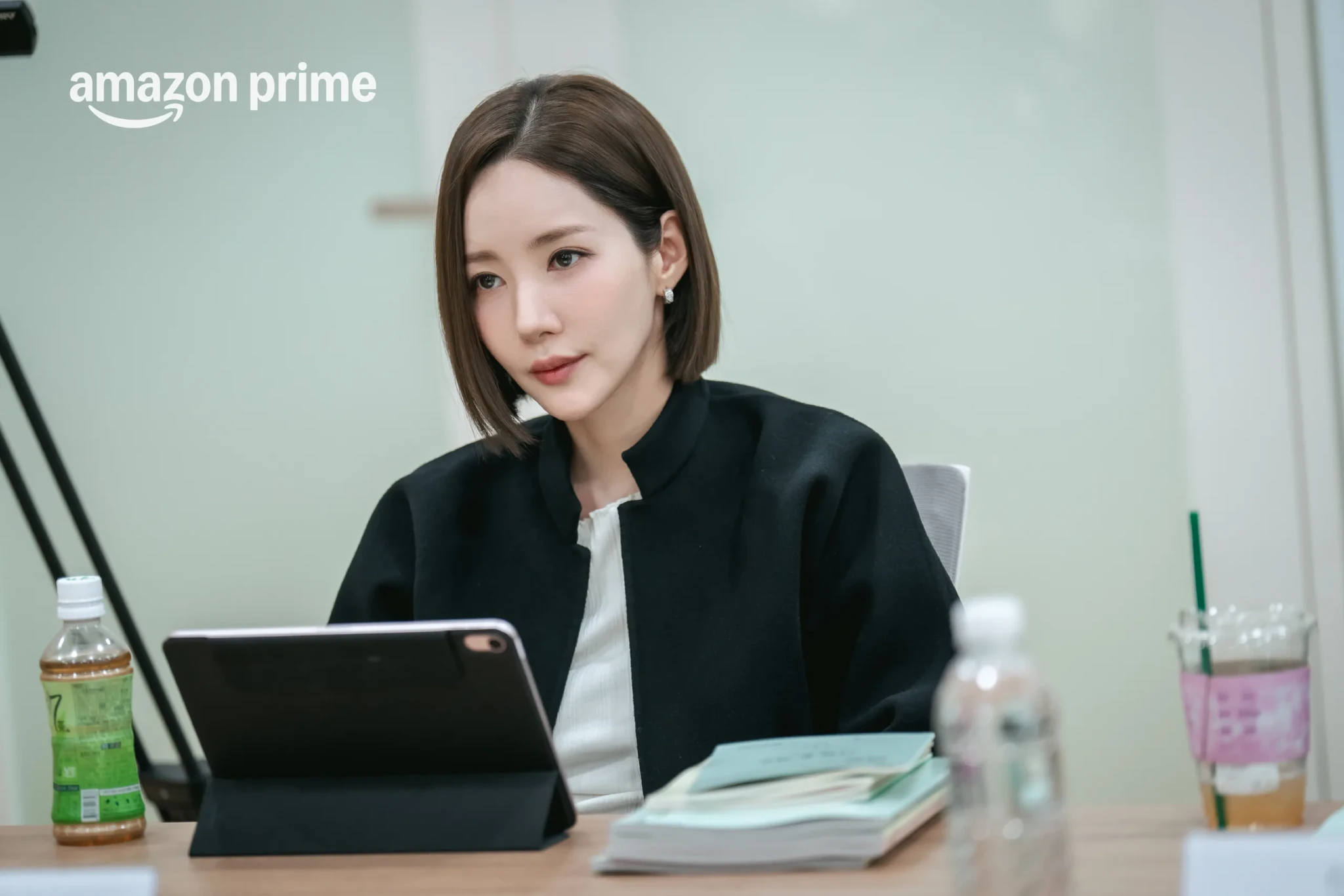 An office scene from Nine to Six showing a woman with a short bob at a desk, looking at a tablet beside stacks of papers and drinks