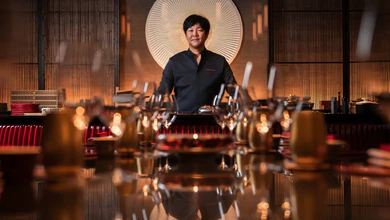 Chef Chen Kentaro standing behind the chef's counter at Shisen Hanten by Chen Kentaro, with glassware on a glossy table in front