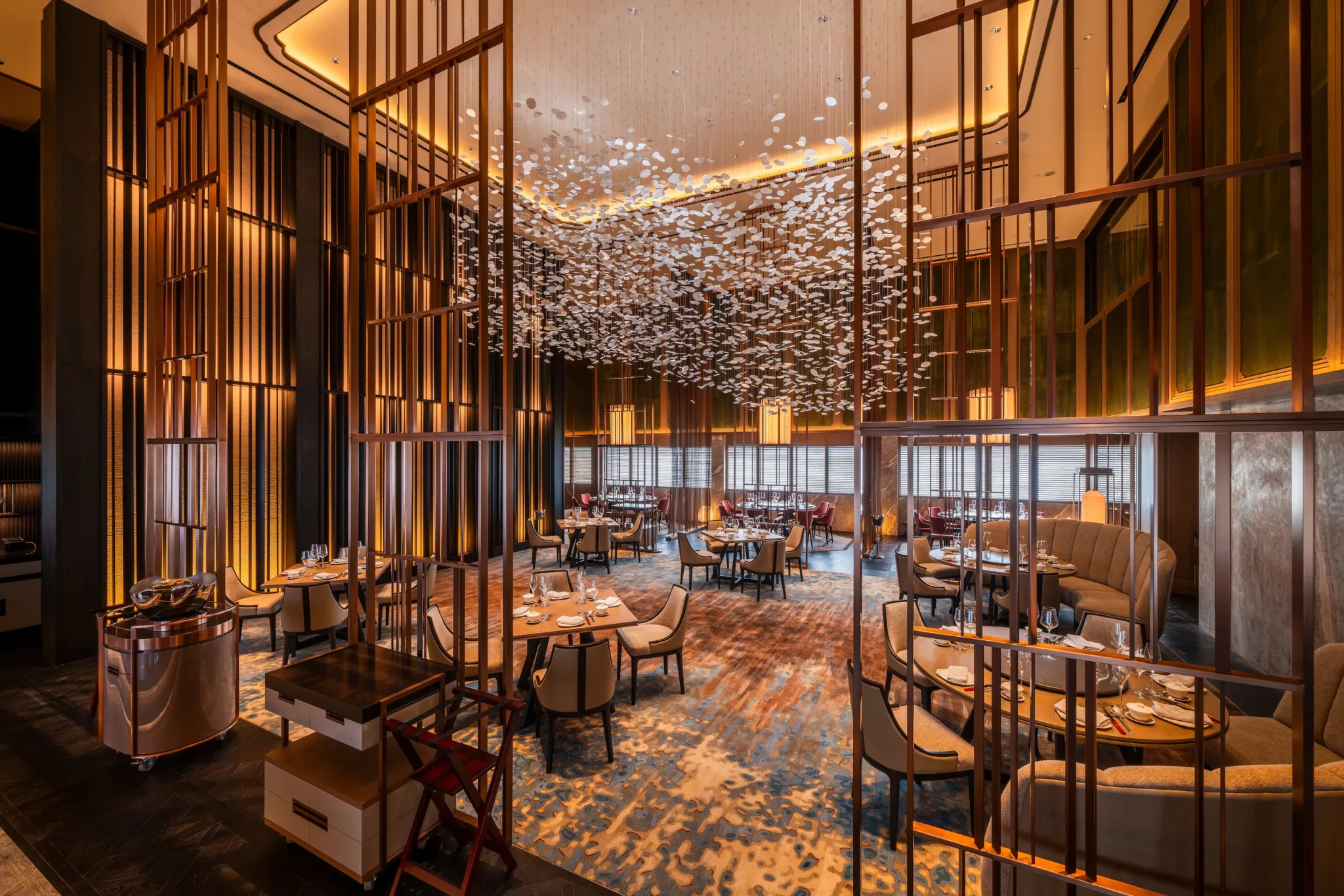 Main dining room with warm lighting, bronze lattice dividers, a white suspended installation overhead, and tables set with glassware.