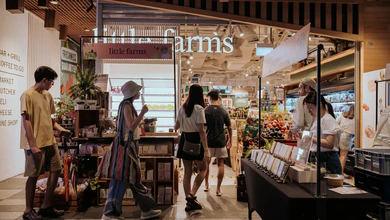 Shoppers walking past the Little Farms entrance, with fresh fruit displays and a produce counter beside the store.