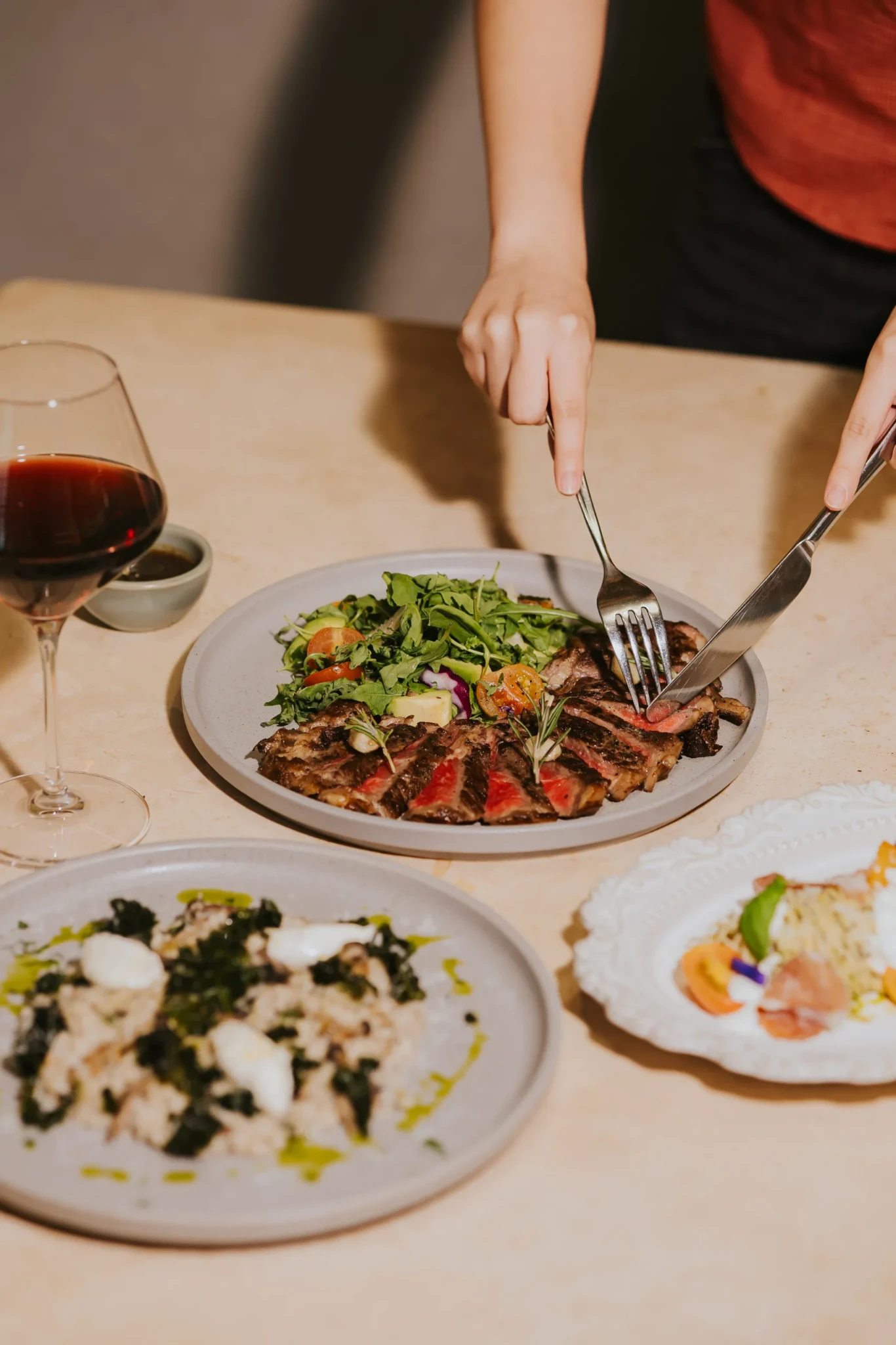 A diner cuts into grilled steak with rosemary and fresh salad, with a glass of red wine on the table